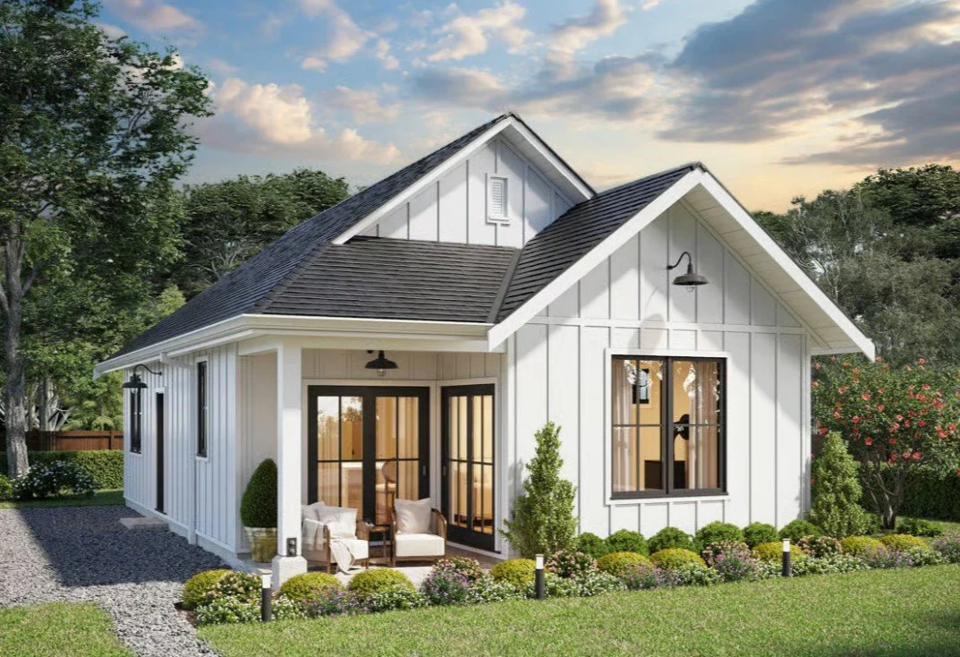 A modern white house with black trim, surrounded by greenery and pink flowers, featuring a small porch with seating, large windows, and a sloped roof.