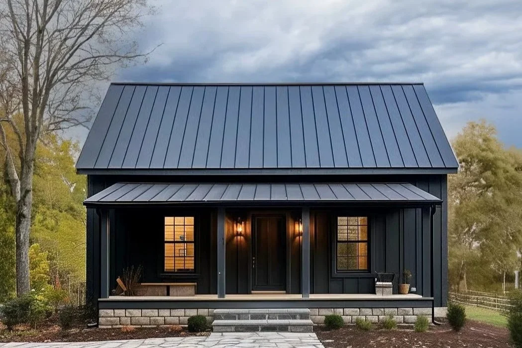 Modern black house with metal roof, front porch with two windows and front door, surrounded by trees and a cloudy sky.