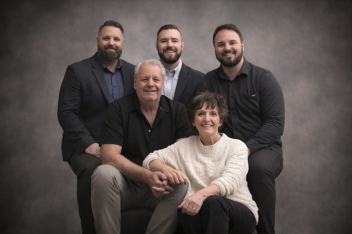 Family portrait of six people, three men and a woman sitting in front, two men standing behind them, all smiling against a plain gray background.