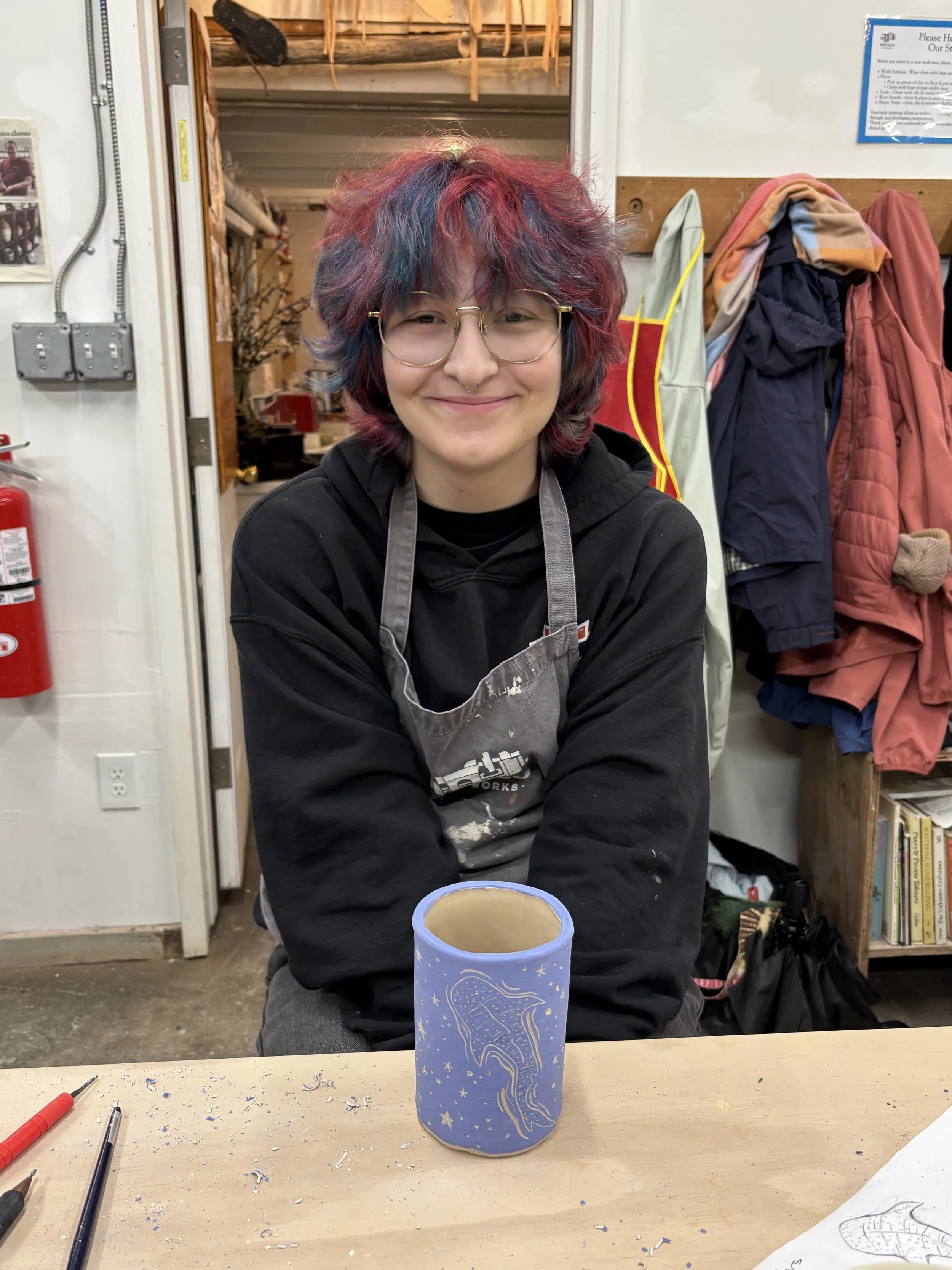 A person with multi-colored, wavy hair and glasses smiling, sitting at a work table with a decorated ceramic cup in front of them, in a workshop or studio setting.