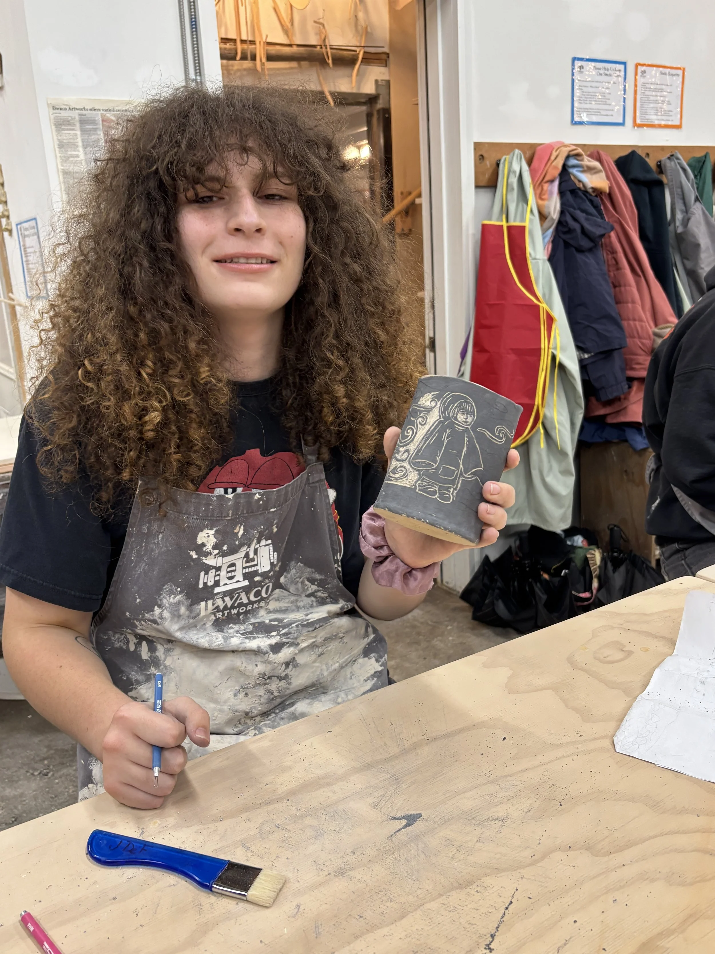 A woman with curly hair and an apron holding a carved piece of stone or wood with a figure etched on it, sitting at a worktable with art supplies in an art studio.