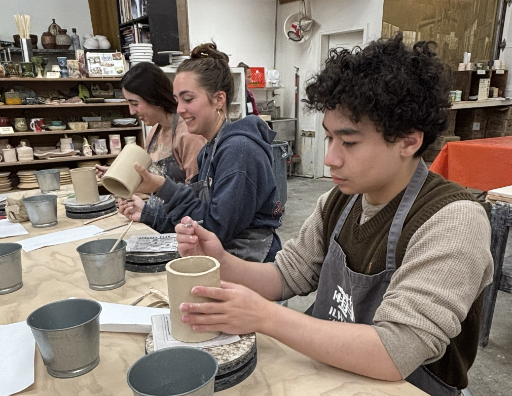 Three young people working on pottery projects at a ceramics studio, shaping and glazing clay mugs.