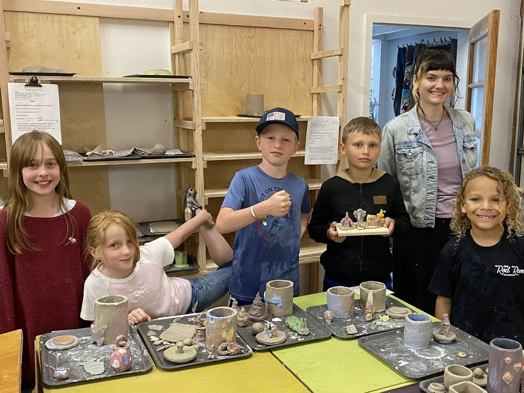 Group of children and an adult woman at a pottery-making workshop, with unfinished ceramic cups, bowls, and figurines on a yellow table in front of them.