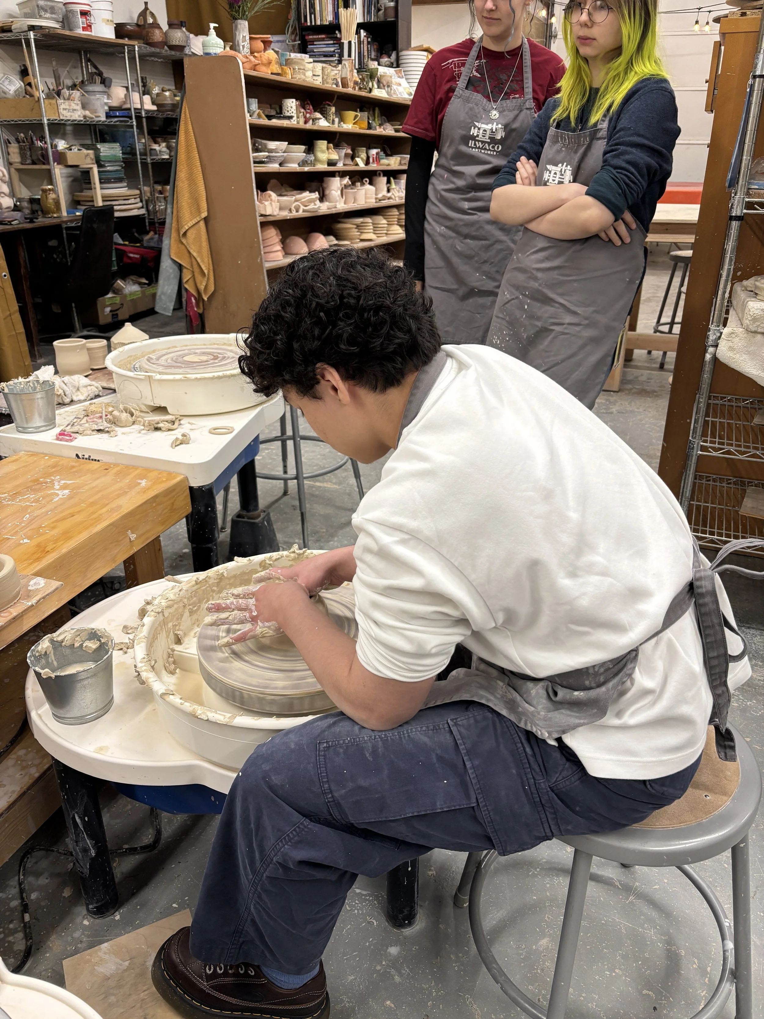 A person shaping clay on a pottery wheel in a pottery studio while two others observe, surrounded by shelves of pottery and supplies.