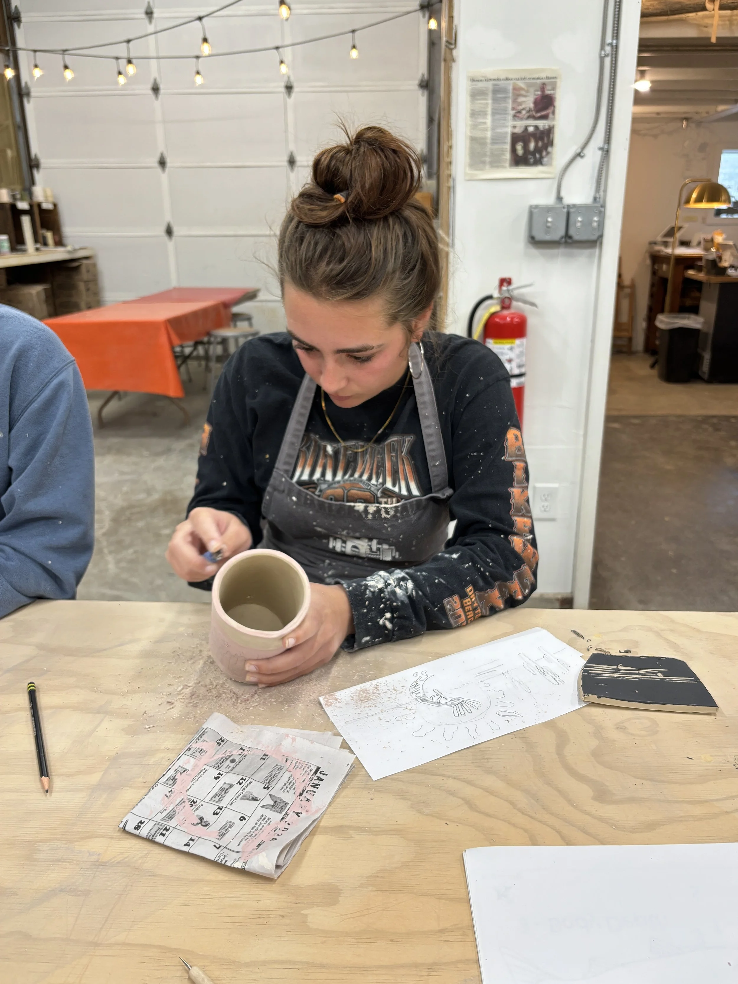 A young woman with a top bun hairstyle, wearing a Detroit Lions sweatshirt and an apron, is working on a ceramic piece at a wooden table, with sketches, a pencil, and a black tool in front of her. She is in a workshop with string lights, a garage door, and a fire extinguisher in the background.