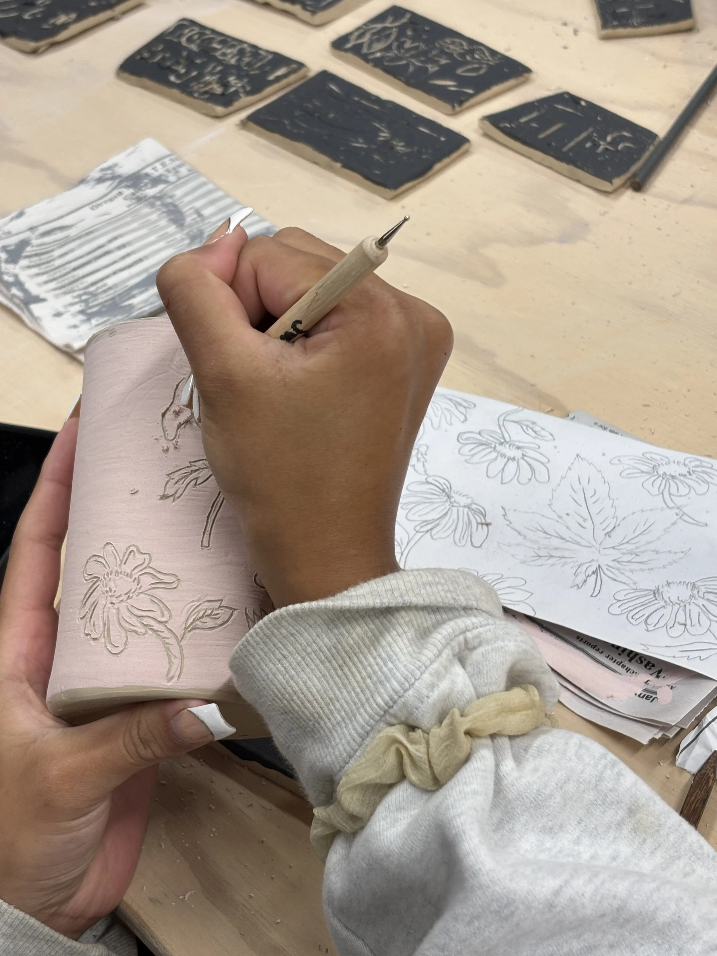 A person sketching floral designs on a pink ceramic piece with a small carving tool, with tiles of black and white designs on the table.