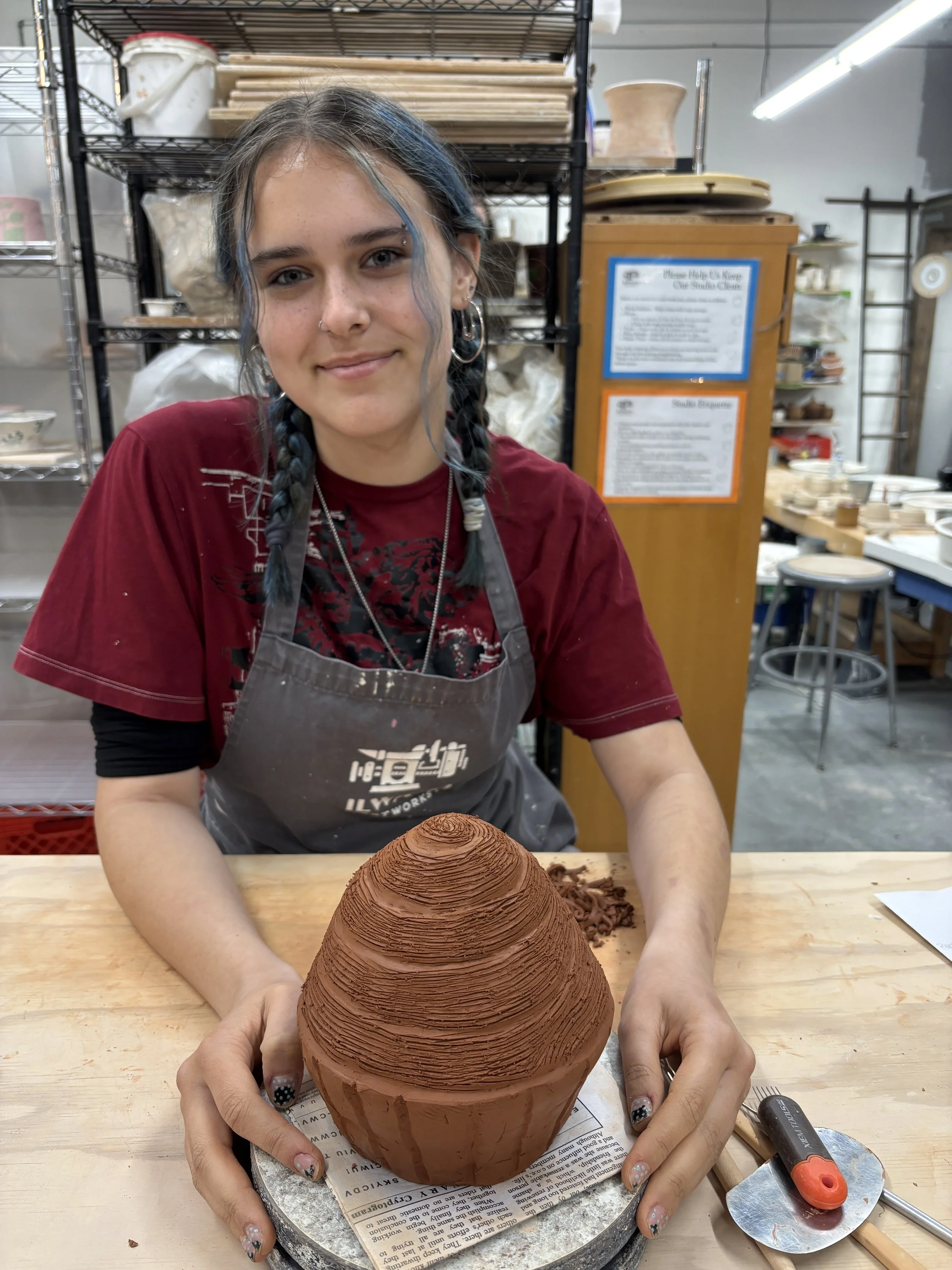 A young woman with braided hair and a nose piercing, smiling at the camera, wearing a maroon T-shirt and an apron, sitting at a worktable with a large, cone-shaped clay sculpture in front of her.