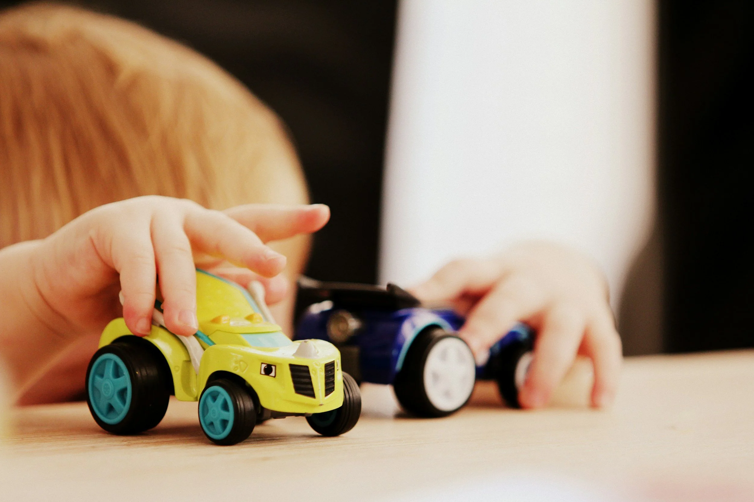Close-up of a child's hand playing with two toy cars on a wooden surface, with the child wearing a brown striped shirt and a person in a black and white outfit in the background.