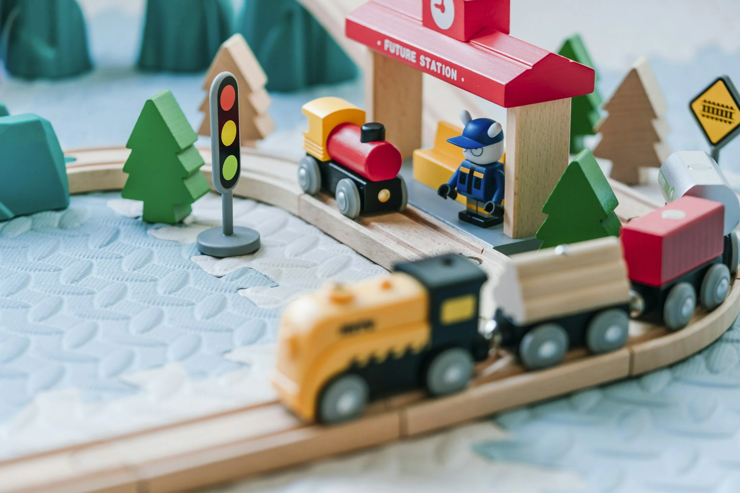 Wooden toy train with yellow locomotive and red cars, a train station with a figure of a train conductor, trees, a traffic light, and a laser crossing sign, all set on a textured blue surface.