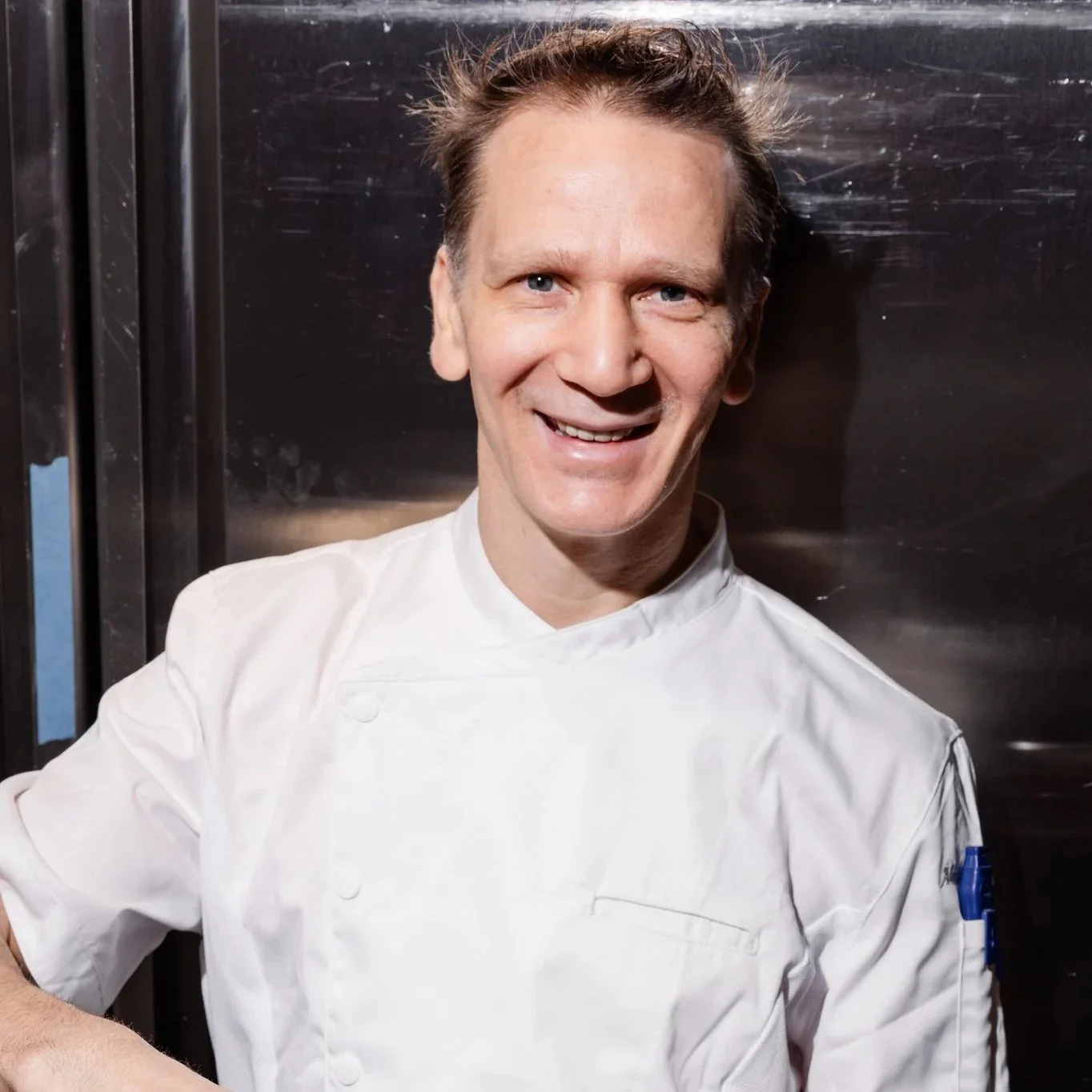 A smiling male chef in a white chef's coat stands in a commercial kitchen.