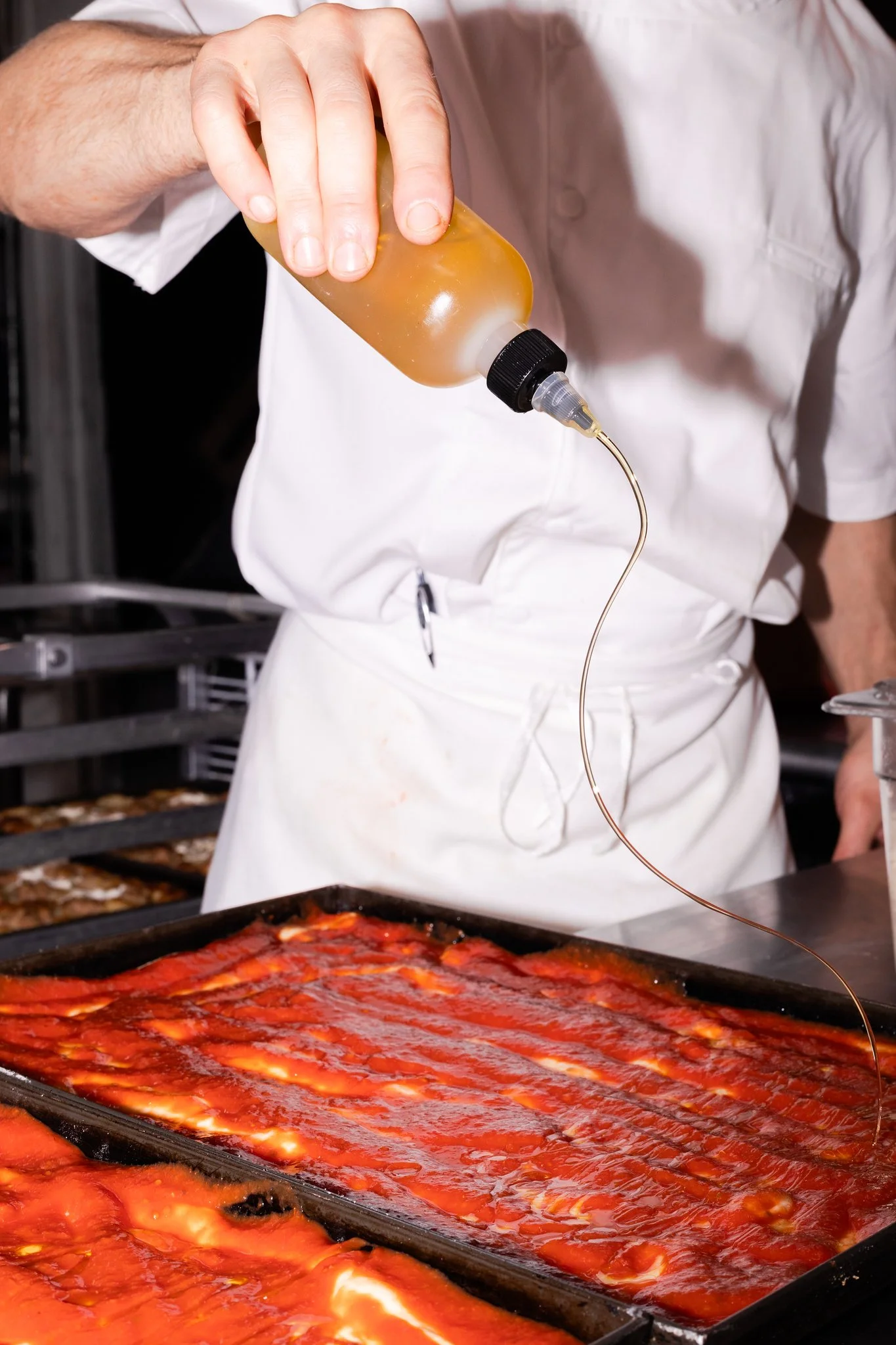 A chef is pouring extra-virgin olive oil from a squeeze bottle onto racks of soon-to-be margherita pizza in a commercial kitchen.