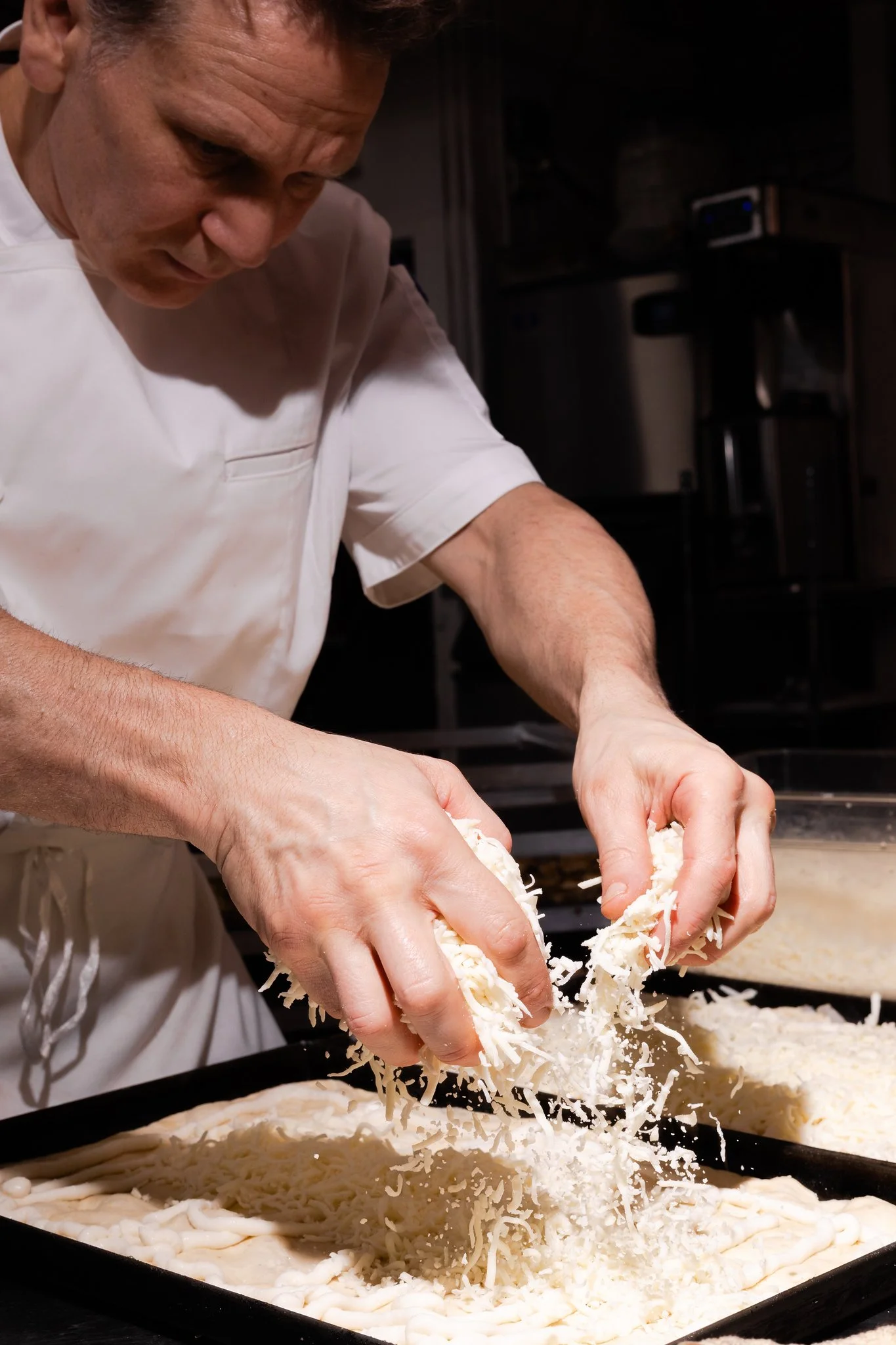A chef in a white uniform preparing a pizza, adding shredded cheese onto the dough.