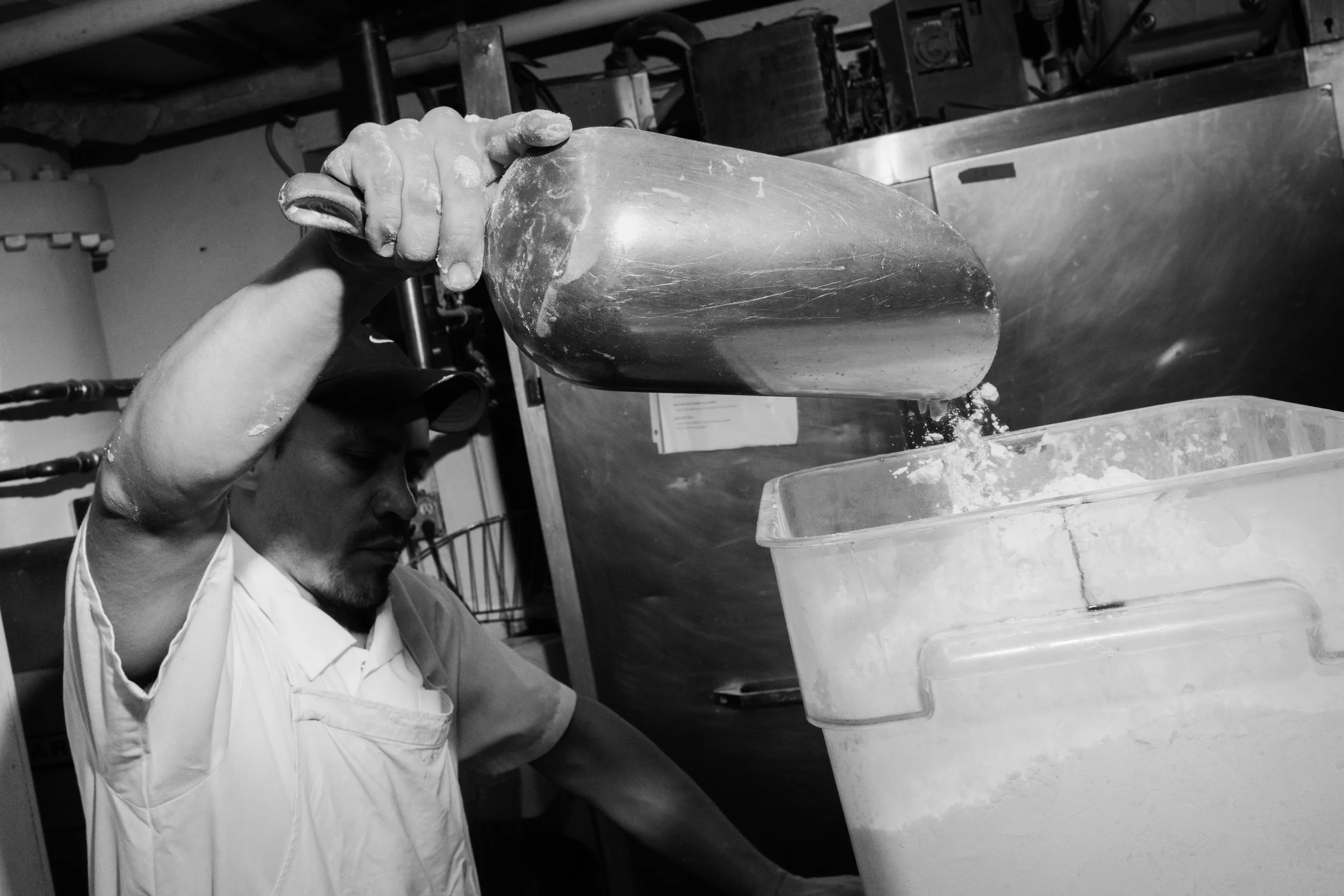 A man wearing a cap and apron is pouring flour mixture from a large metal scoop into a container in a commercial kitchen and bakery.