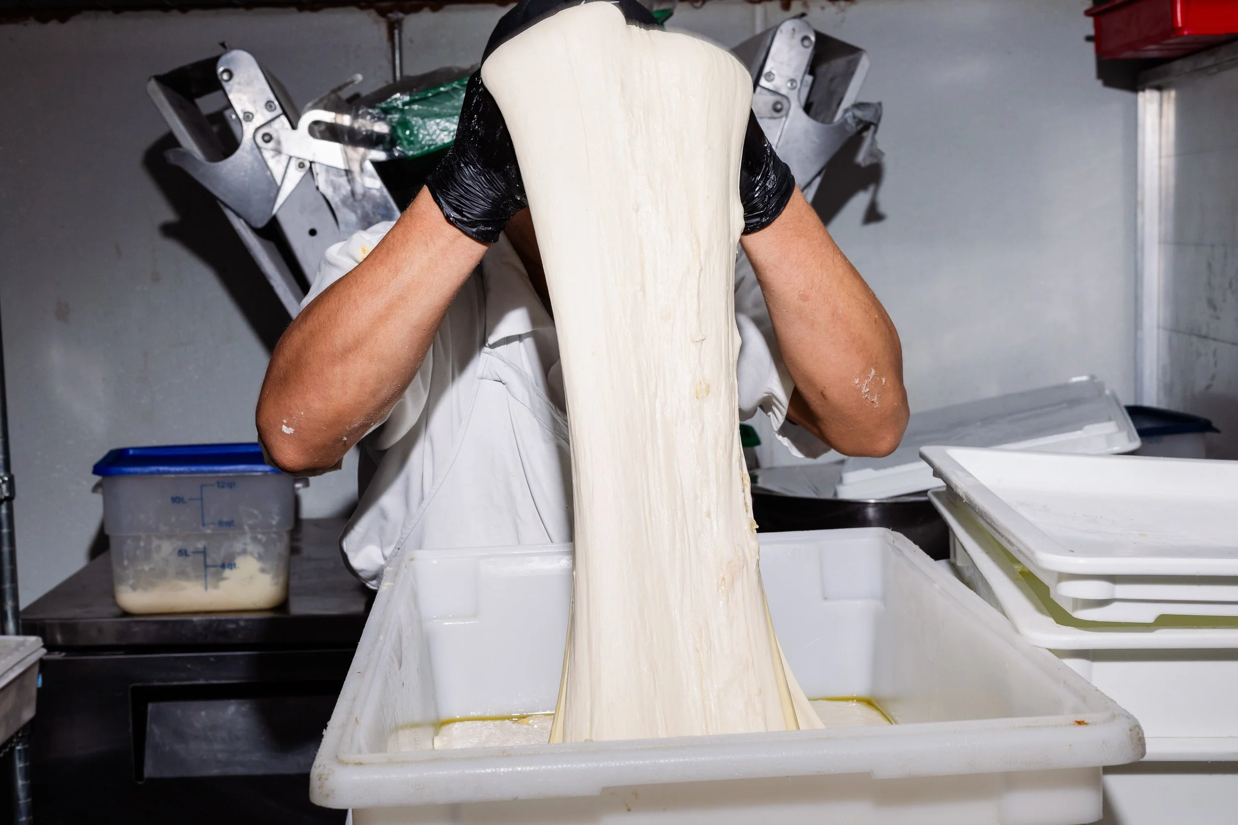 A person pizza dough in a professional kitchen with white containers and equipment around.