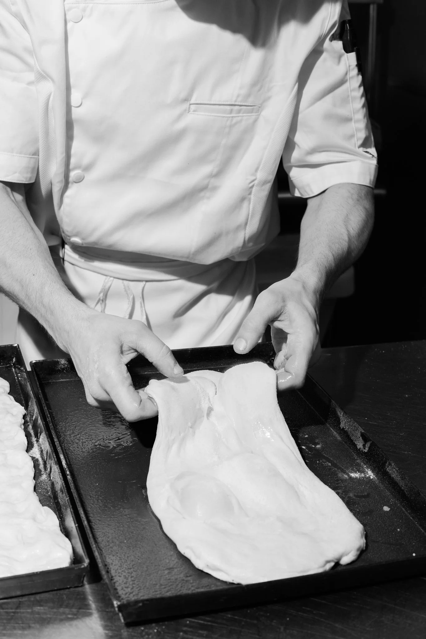 Chef shaping pizza dough on a black baking sheet in a professional kitchen.