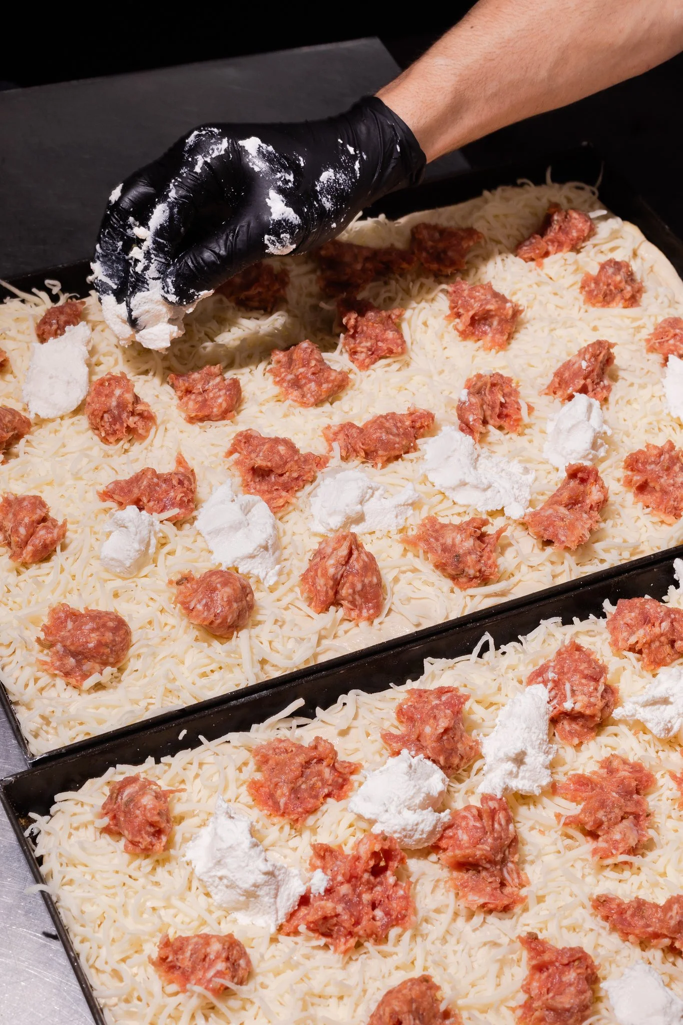 Chef setting ricotta cheese over uncooked roman-style pizza tray topped with shredded cheese, raw sausage, and dollops of ricotta cheese.