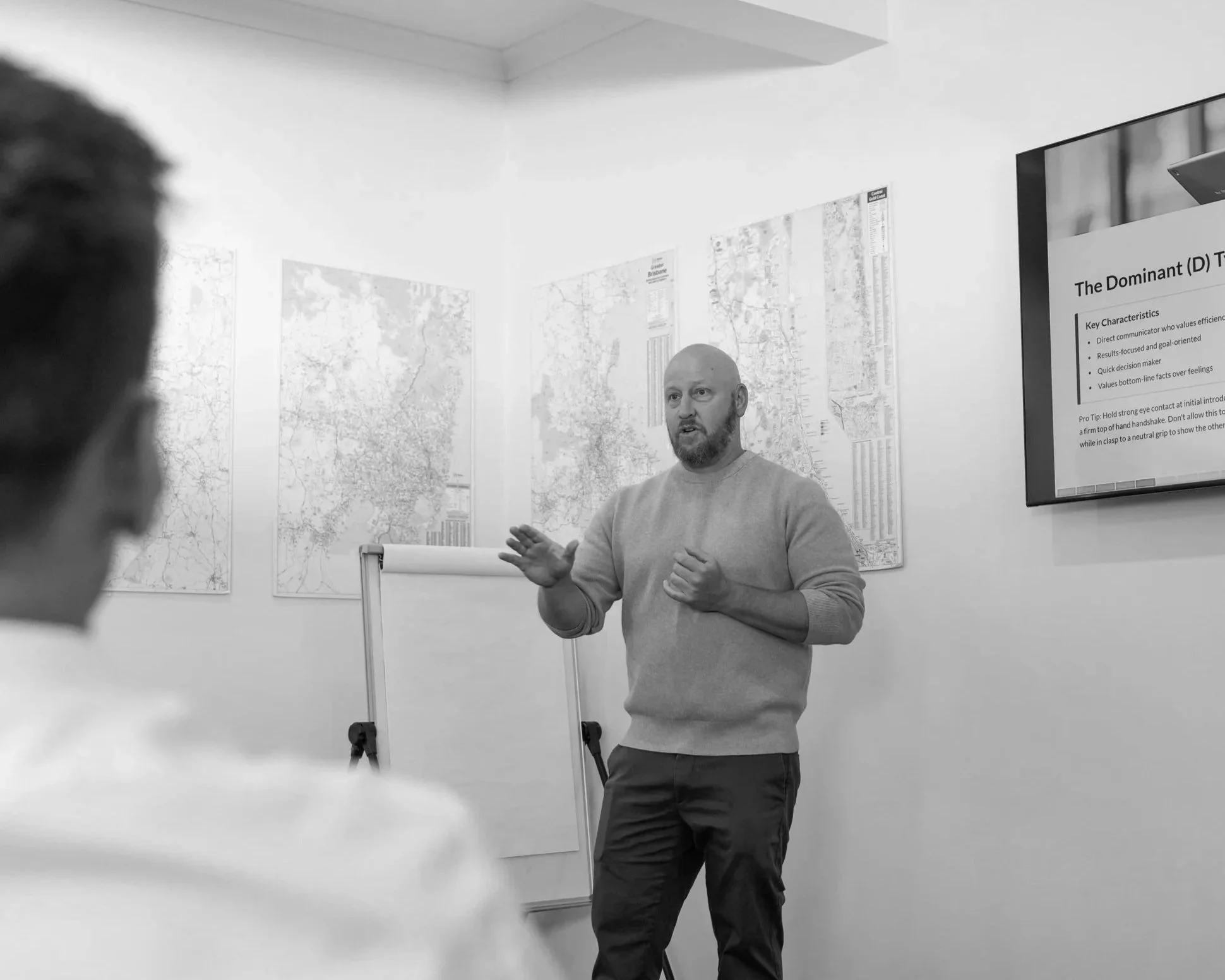 A man giving a presentation to an audience in a room with maps on the wall and a presentation slide on a screen. The man is gesturing with his hands, and there are two people in the audience.