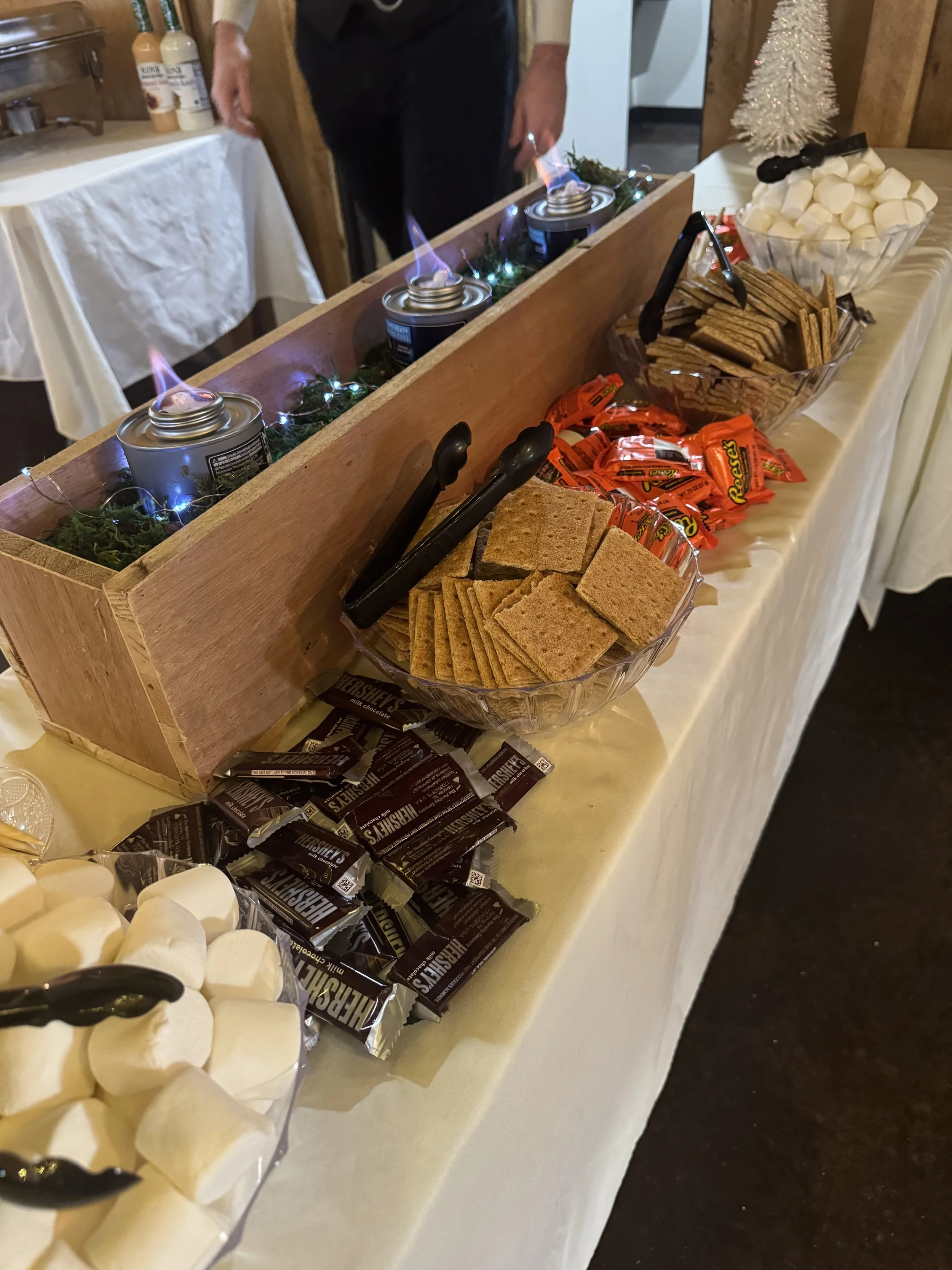 A dessert and snack table featuring marshmallows, Hershey's chocolate bars, graham crackers, Reese's miniatures, and marshmallow fluff. There are three small cans with flames on top, decorated with greenery and string lights.