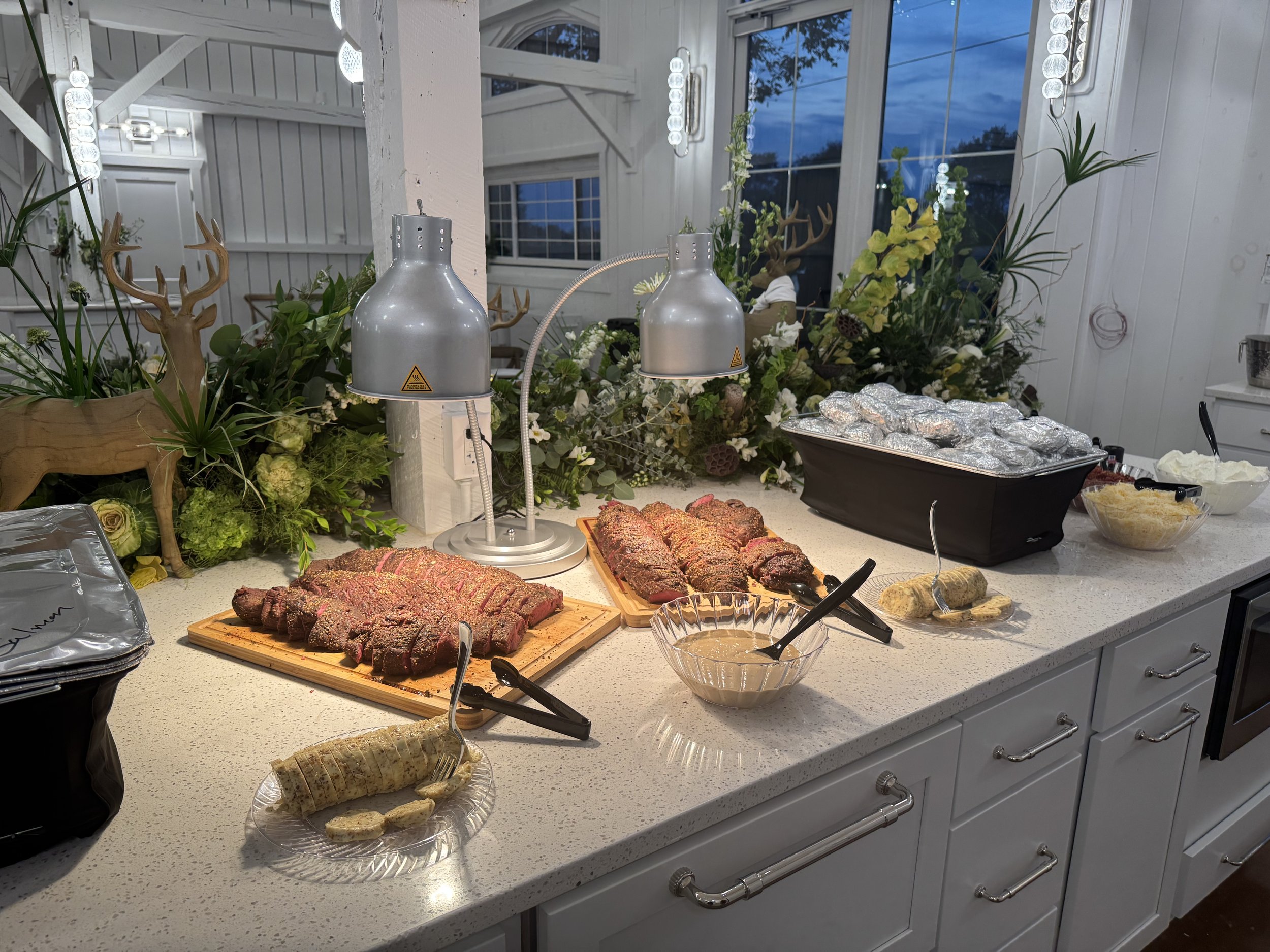 A buffet-style spread with sliced cooked meat, foil-wrapped baked potatoes, dipping sauce, and various side dishes on a white countertop. Decor includes floral arrangements and reindeer figurines.