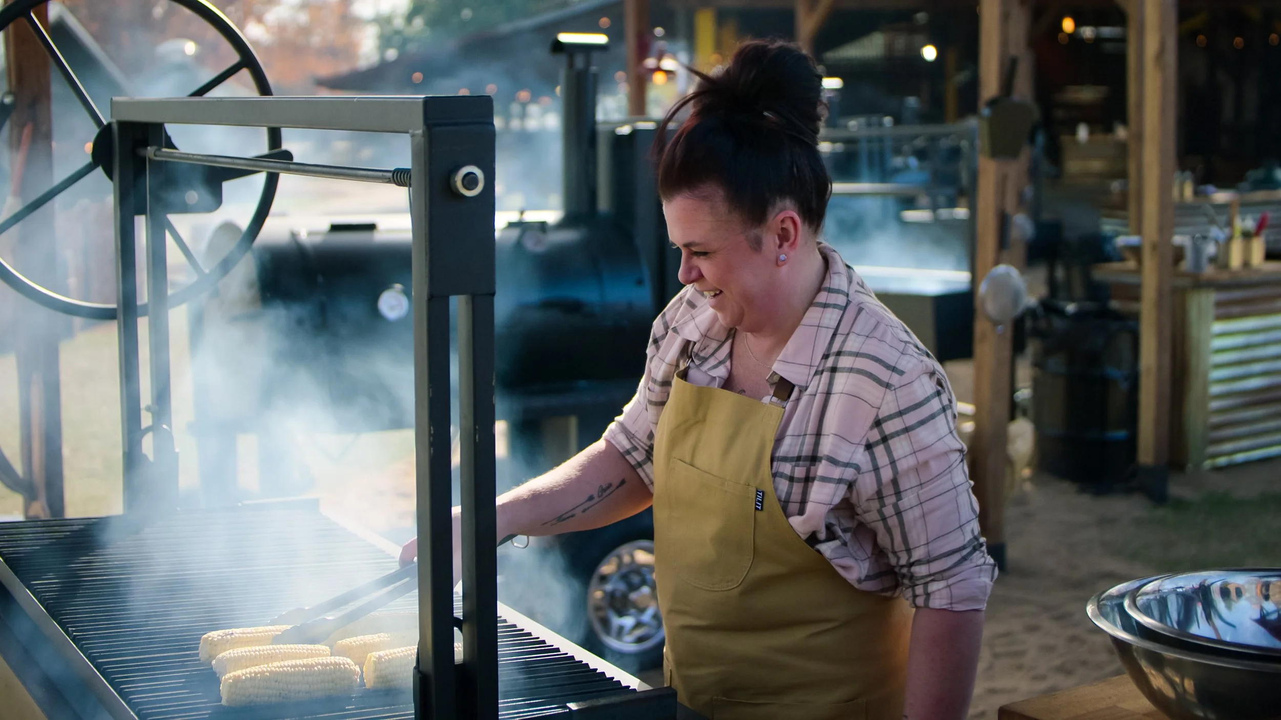 A woman grilling corn on an outdoor barbecue grill, smiling and enjoying cooking.