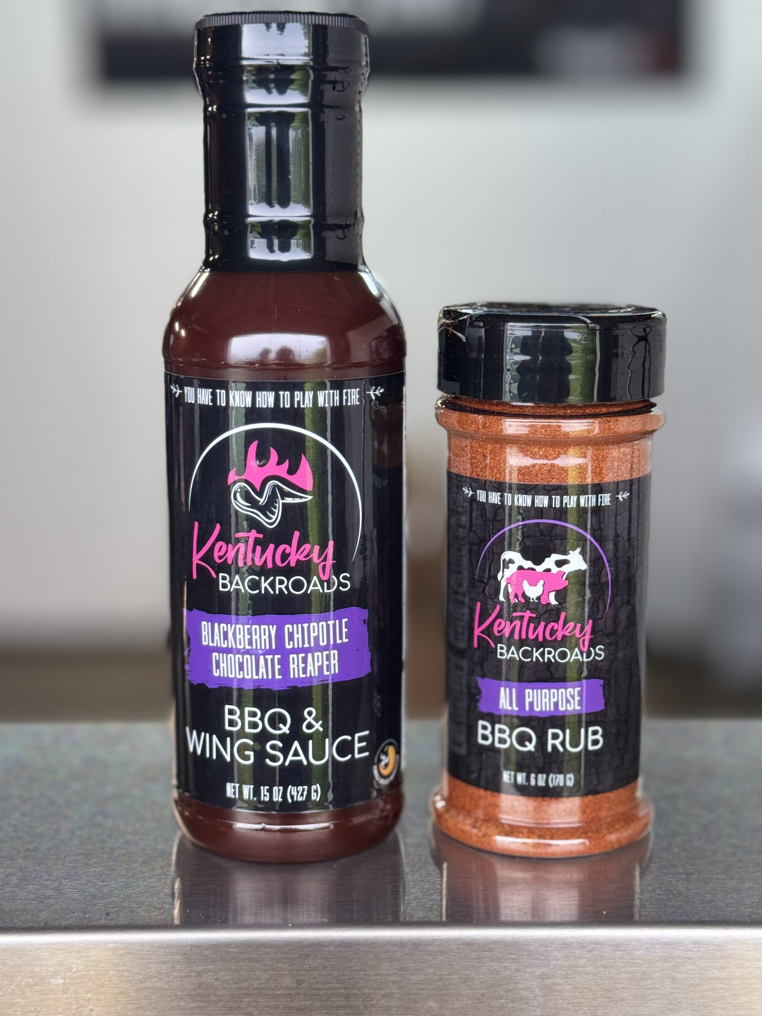 Two bottles of Kentucky Backroads barbecue sauces; one is barbecue and wing sauce, the other is all-purpose barbecue rub, both with black caps and labels with black backgrounds, purple accents, and a pig logo.