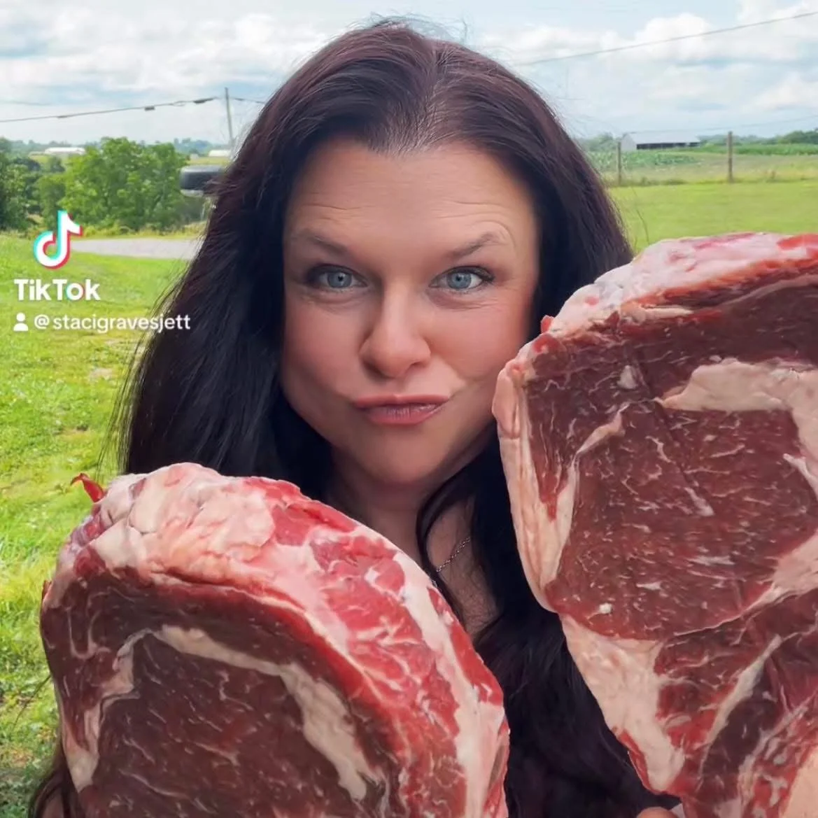 Woman with long dark hair and blue eyes holding large cuts of raw beef in an outdoor field.