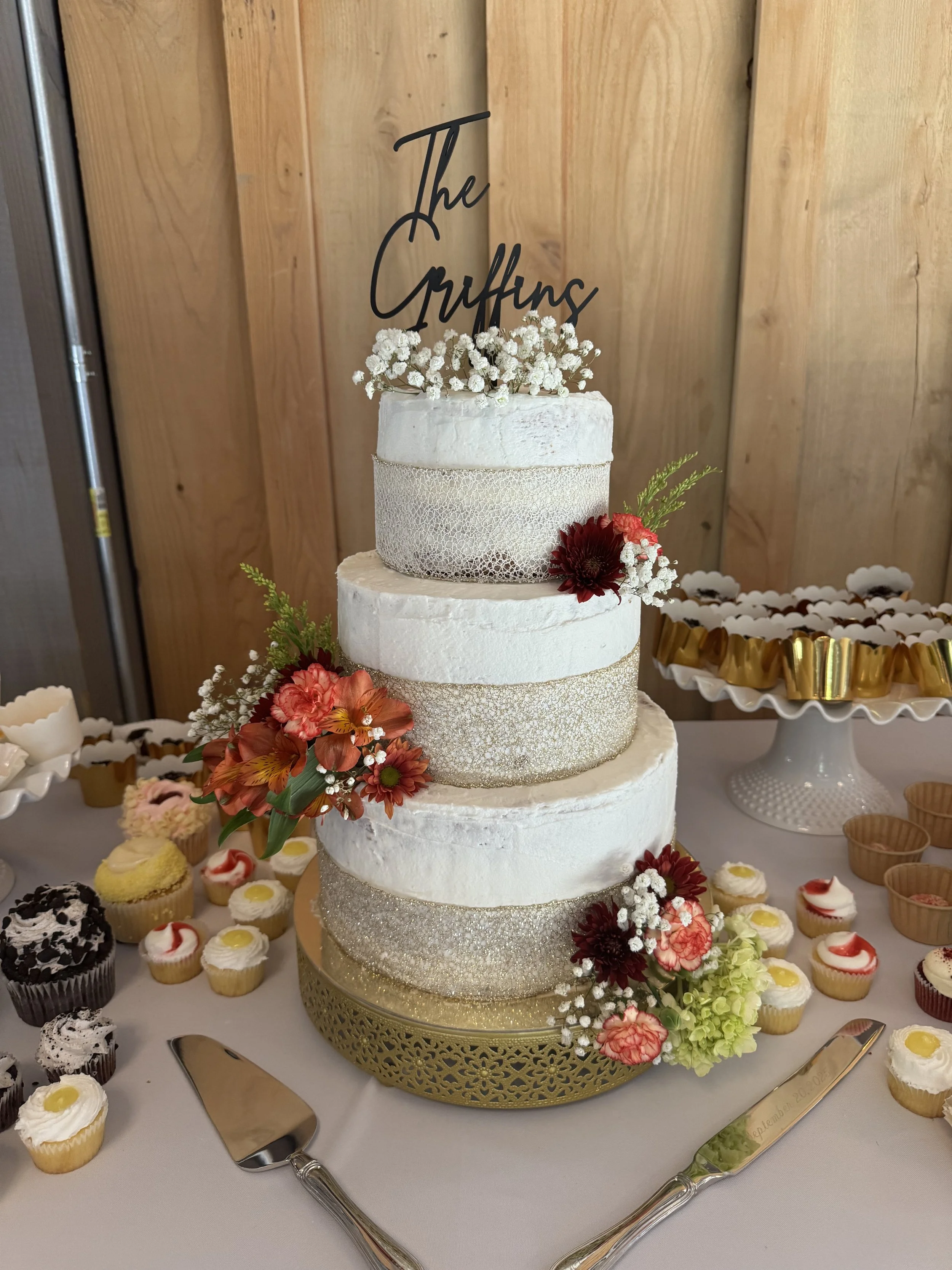 Three-tiered wedding cake decorated with white and gold textured bands, topped with white baby's breath flowers and a 'The Griffins' cake topper. The cake is surrounded by colorful flowers and desserts on a table.