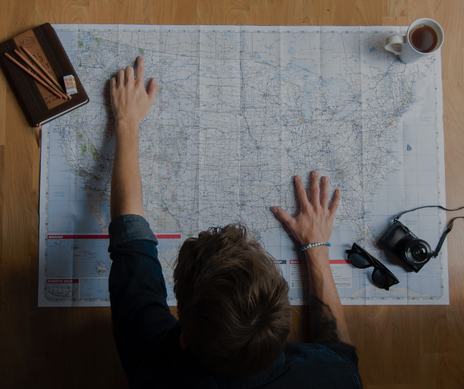 Person planning a trip with a map of the United States, a camera, sunglasses, a cup of tea or coffee, and travel accessories on a wooden table.