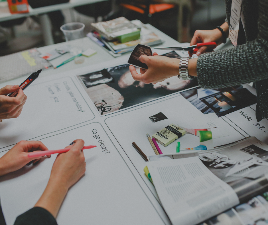 People reviewing photographs and documents on a cluttered table at a workshop or meeting.
