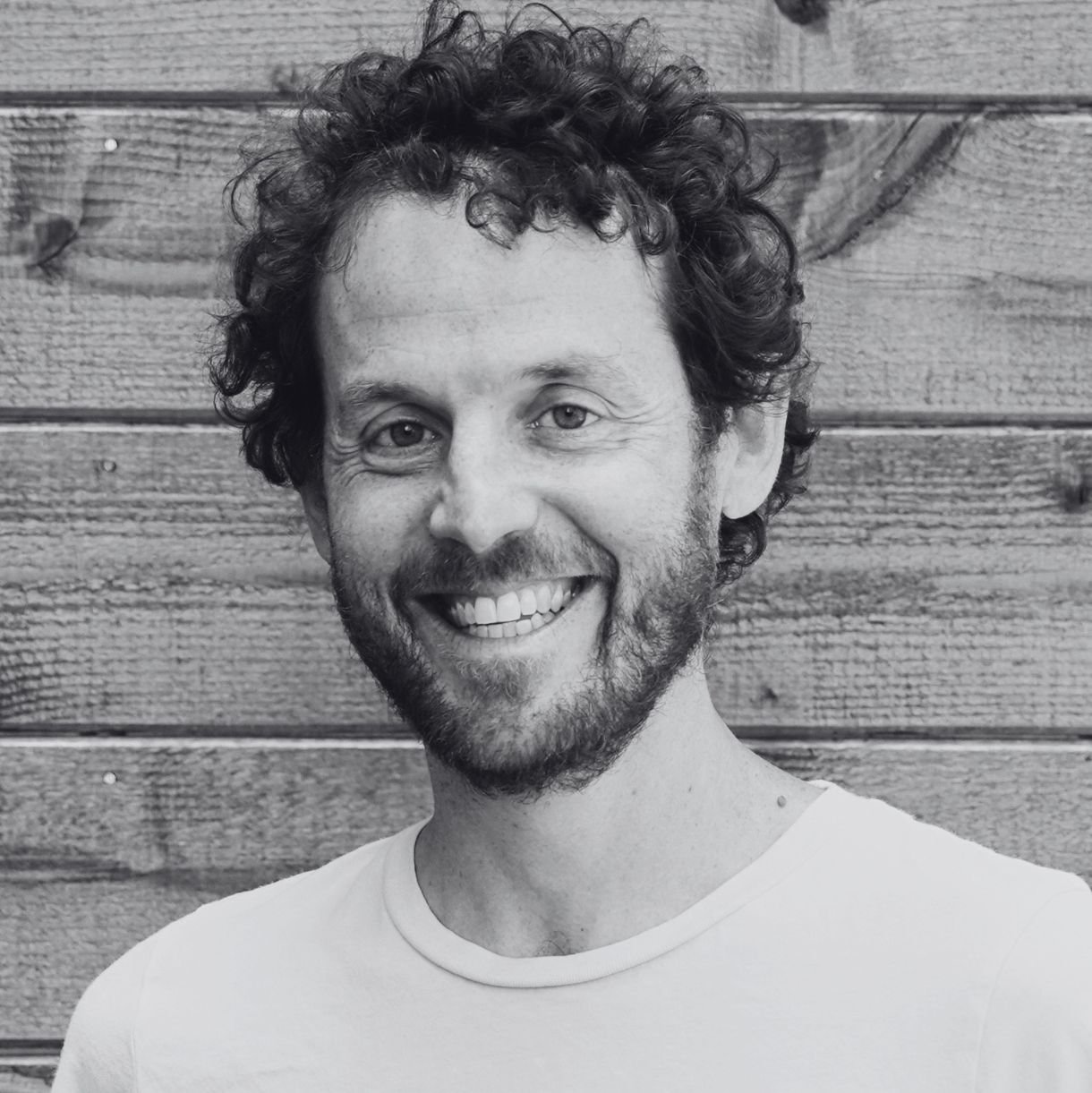 A man with curly hair and a beard smiling in front of a wooden wall.