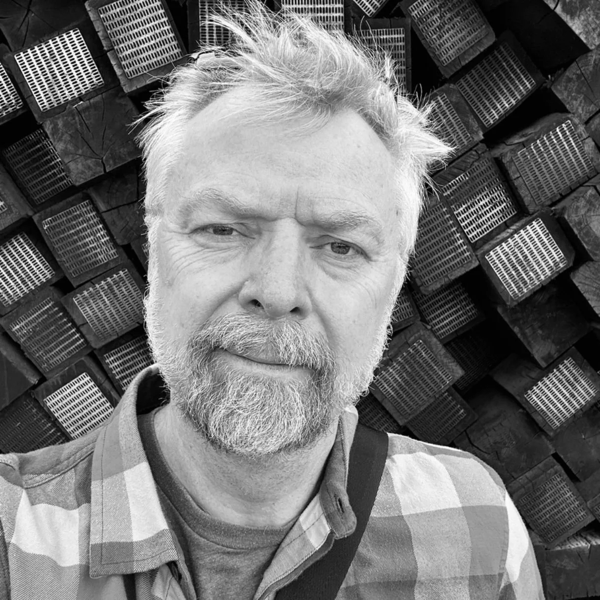 A black-and-white photo of a middle-aged man with a beard and mustache, wearing a checkered shirt, standing in front of stacked firewood.