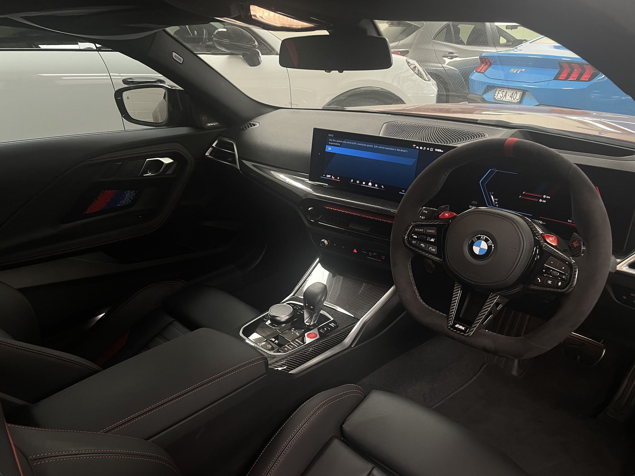 Interior of a BMW sports car with a digital dashboard, a multifunction steering wheel, and a central touchscreen display, parked in a garage with other vehicles visible through the windshield.