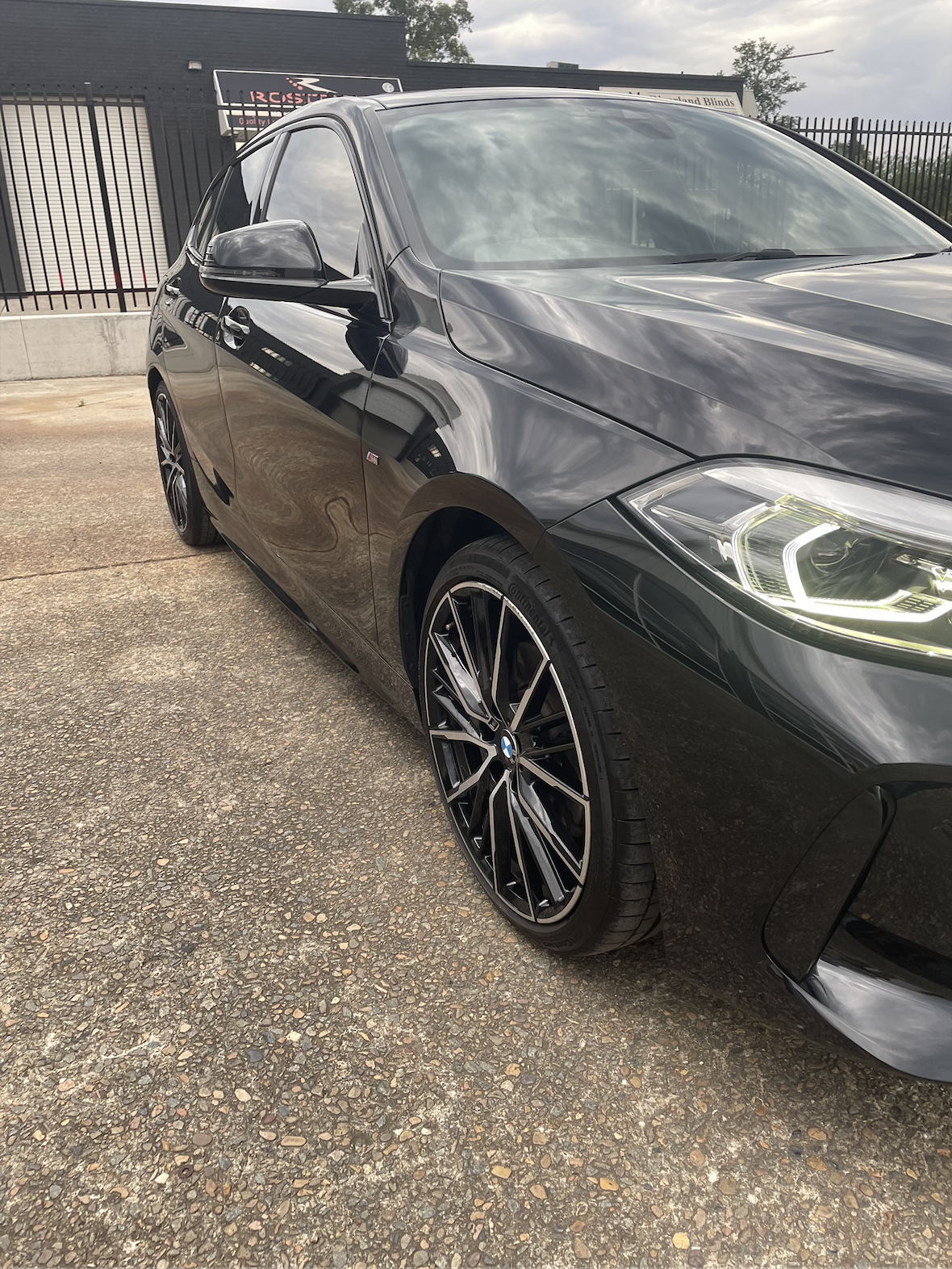 Black BMW car parked on a gravel lot with a black fence and building in background.