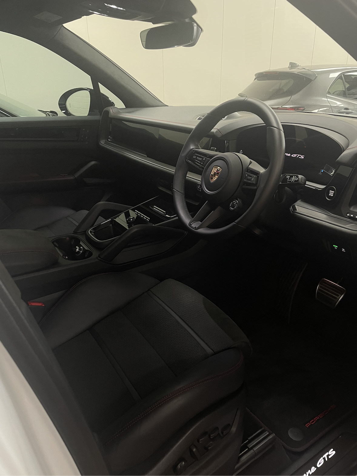 Interior of a Porsche vehicle, showing the steering wheel, dashboard, center console, and front seat, with another car visible outside the window.