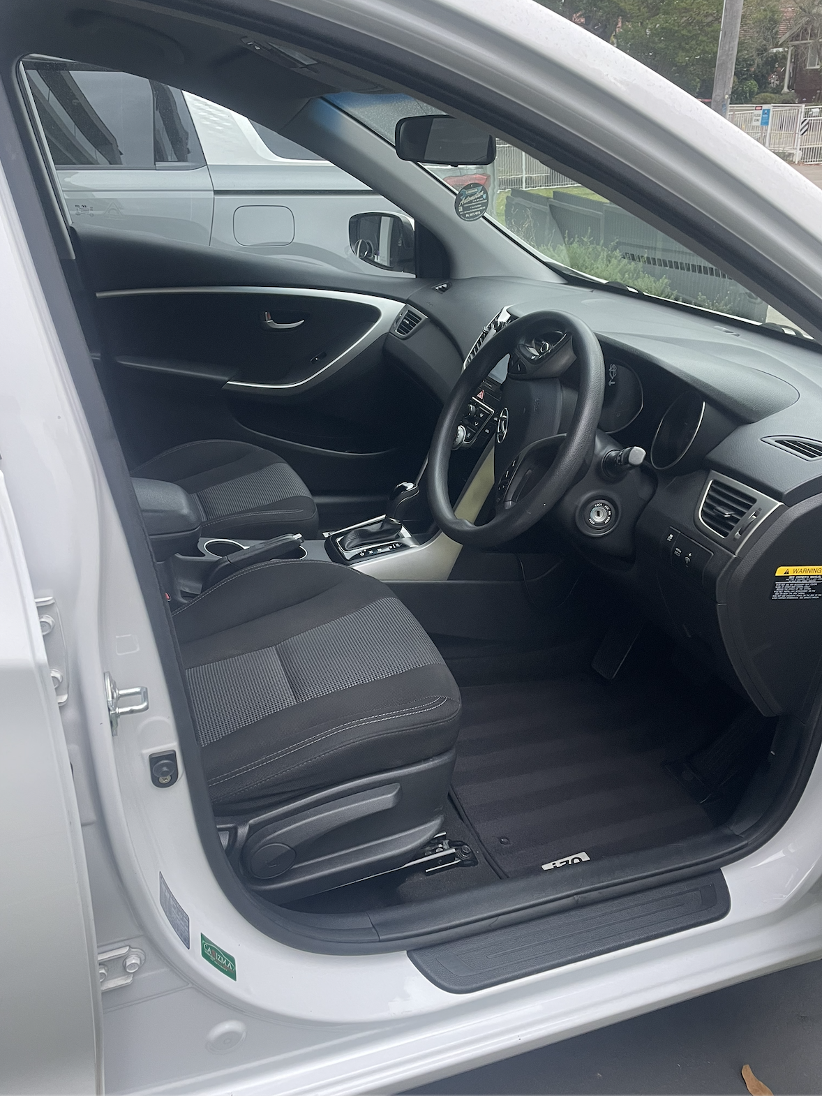 Interior of a white car showing the driver's seat, steering wheel, dashboard, and part of the passenger seat, with an outside view of a parked vehicle and a fence.