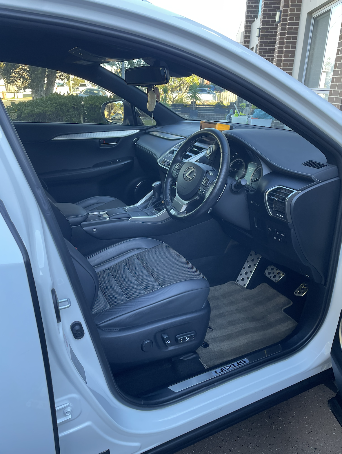 Interior of a white Lexus car showing black seats, steering wheel, dashboard, and pedals, with a view of trees and houses outside.