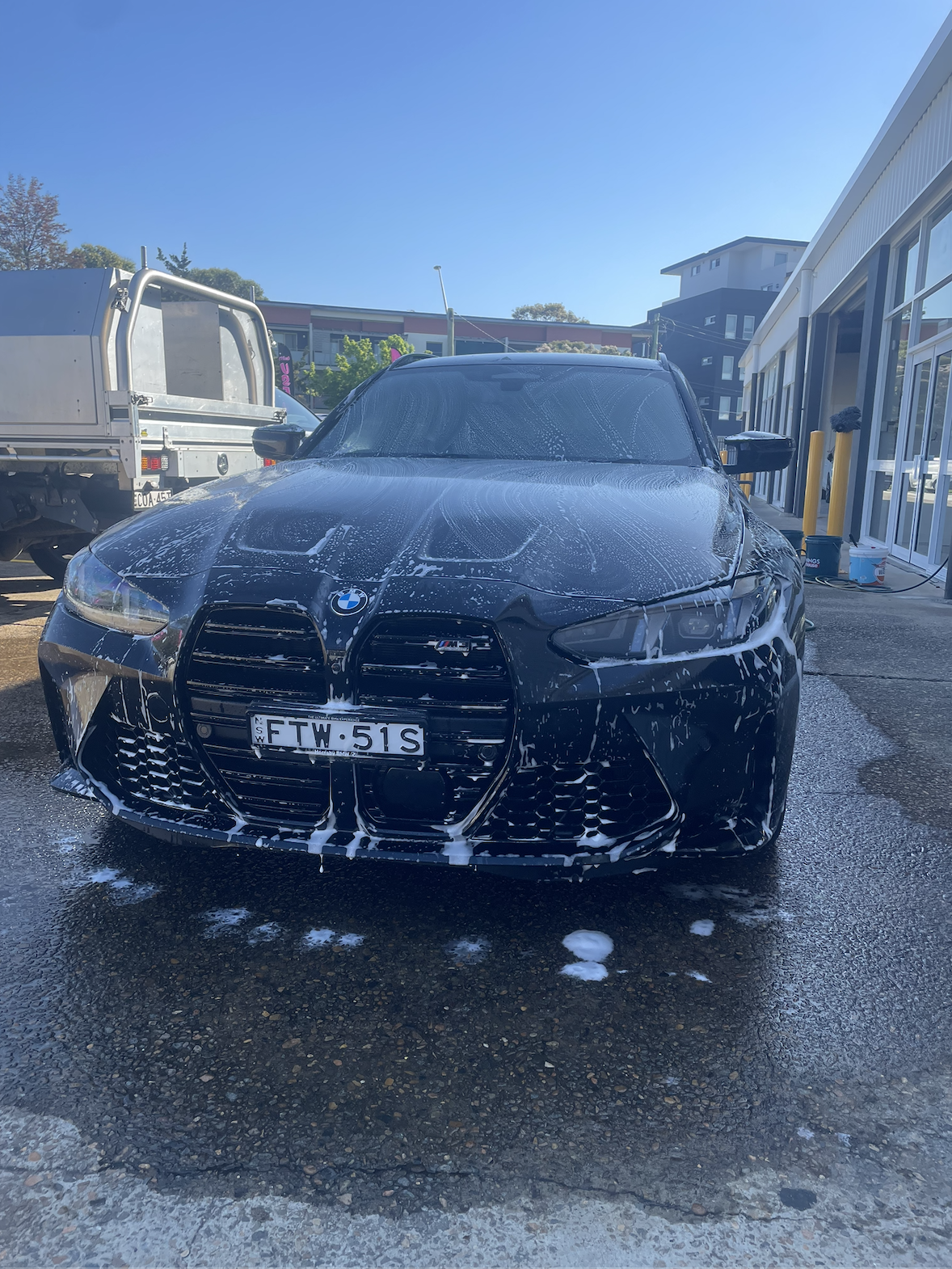 Black BMW M4 with soap suds on front and windshield, in a car wash station, with soap foam on the ground, behind a truck, and buildings in the background.