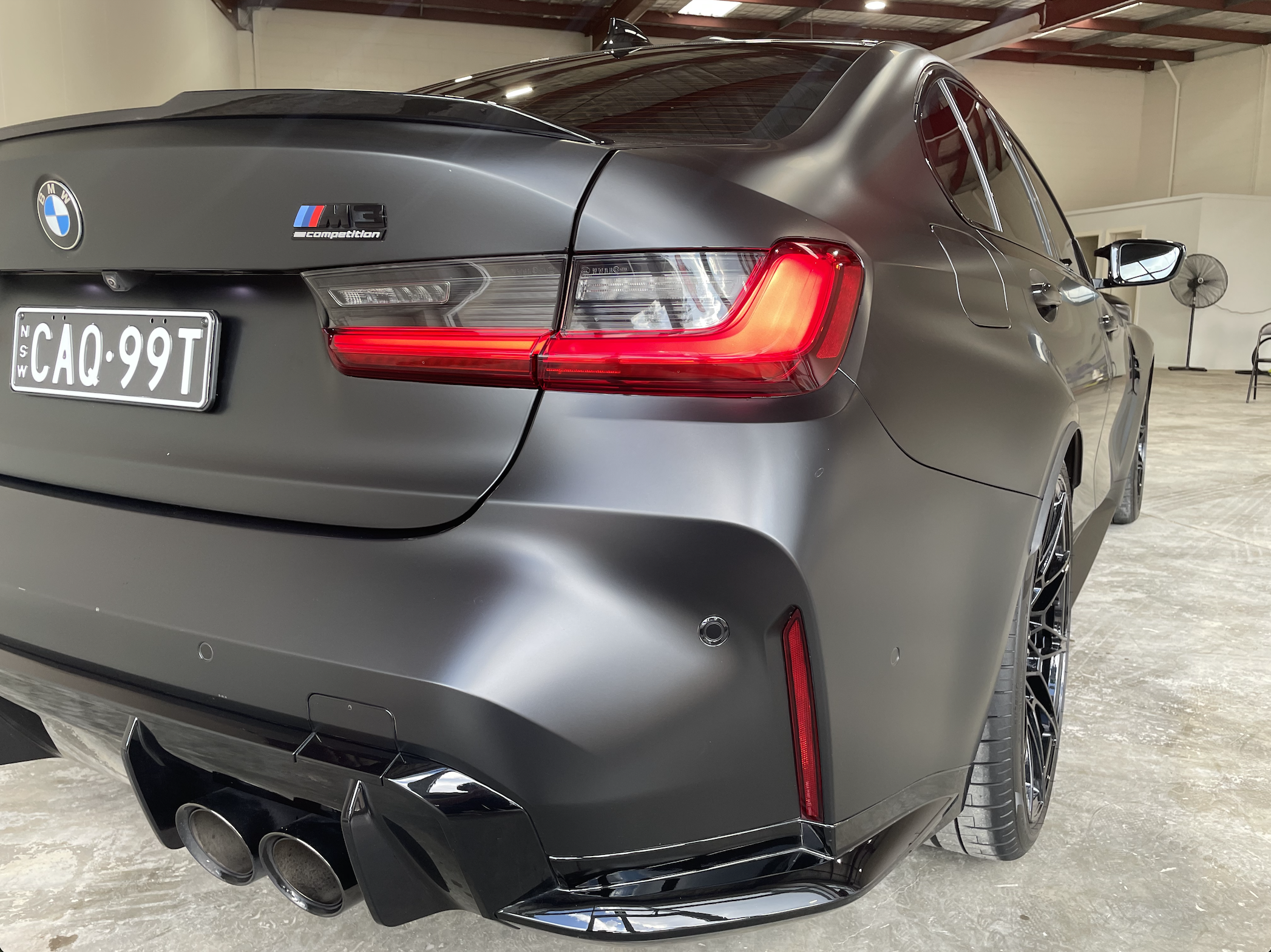 A matte black BMW M3 car parked indoors, displaying the rear with taillights, quad exhaust pipes, and M3 competition badge.