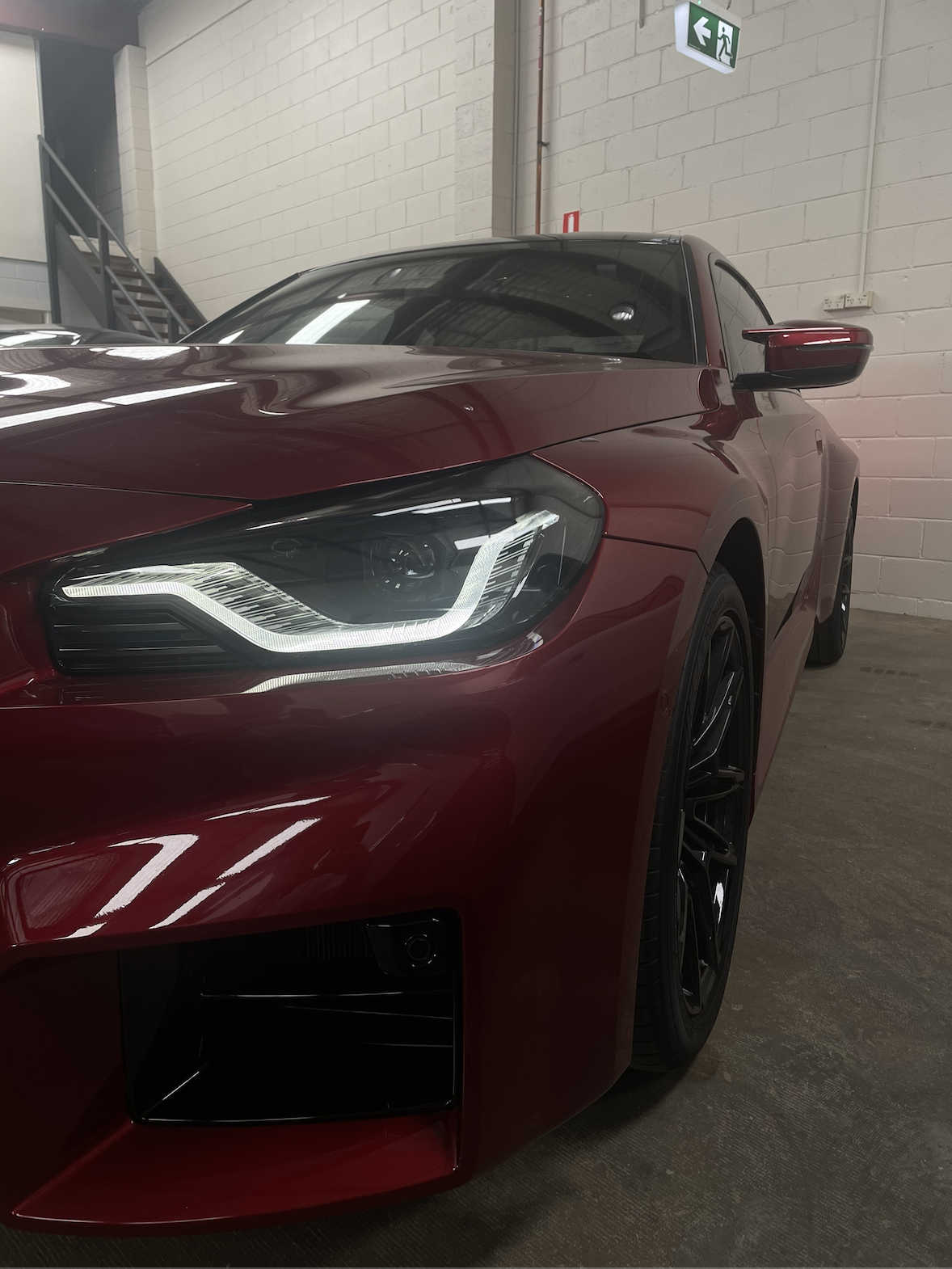 Close-up of the front left side of a red sports car with sleek headlights parked indoors against a white brick wall.
