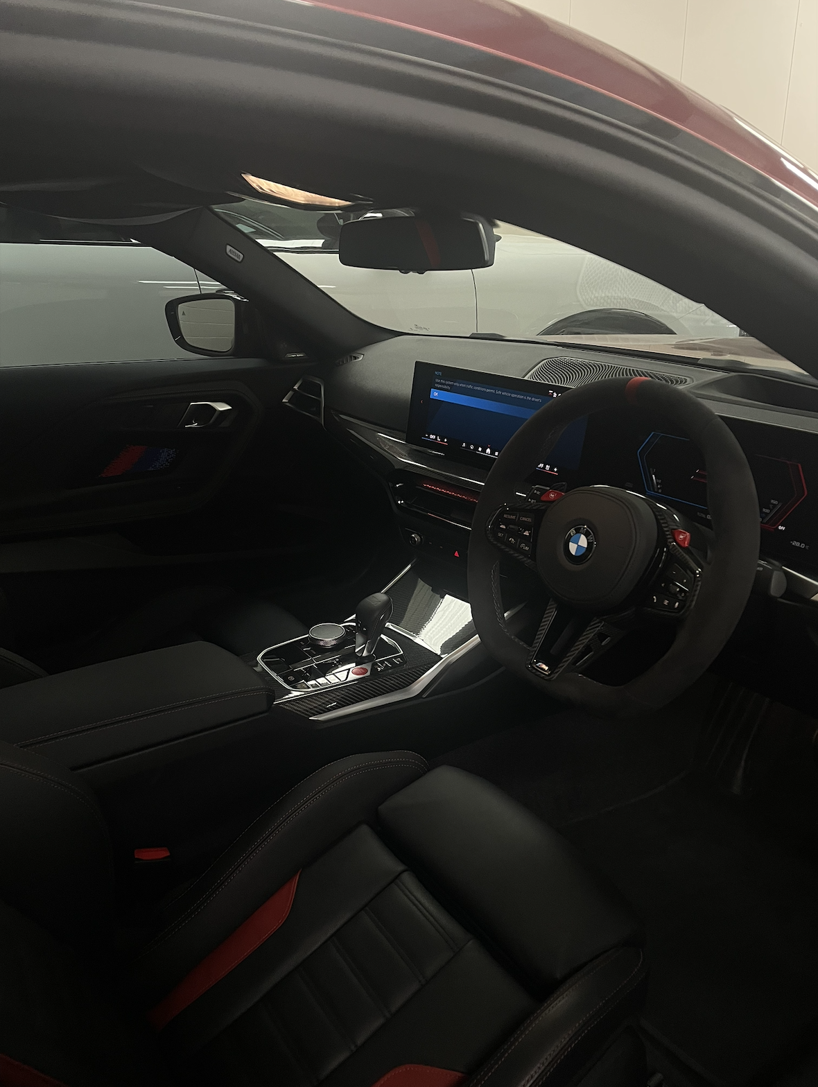 Interior of a BMW car showing the dashboard, steering wheel, gear shift, and infotainment screen. The interior features black leather with red accents.