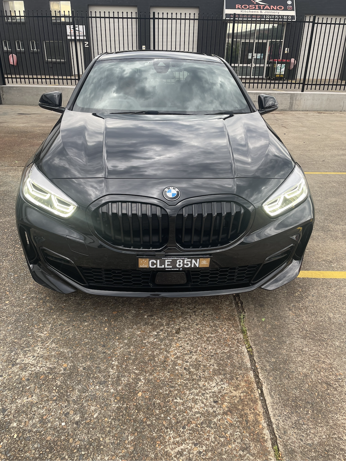 Front view of a black BMW car parked on a concrete surface in front of a black fence, with buildings and signage in the background.