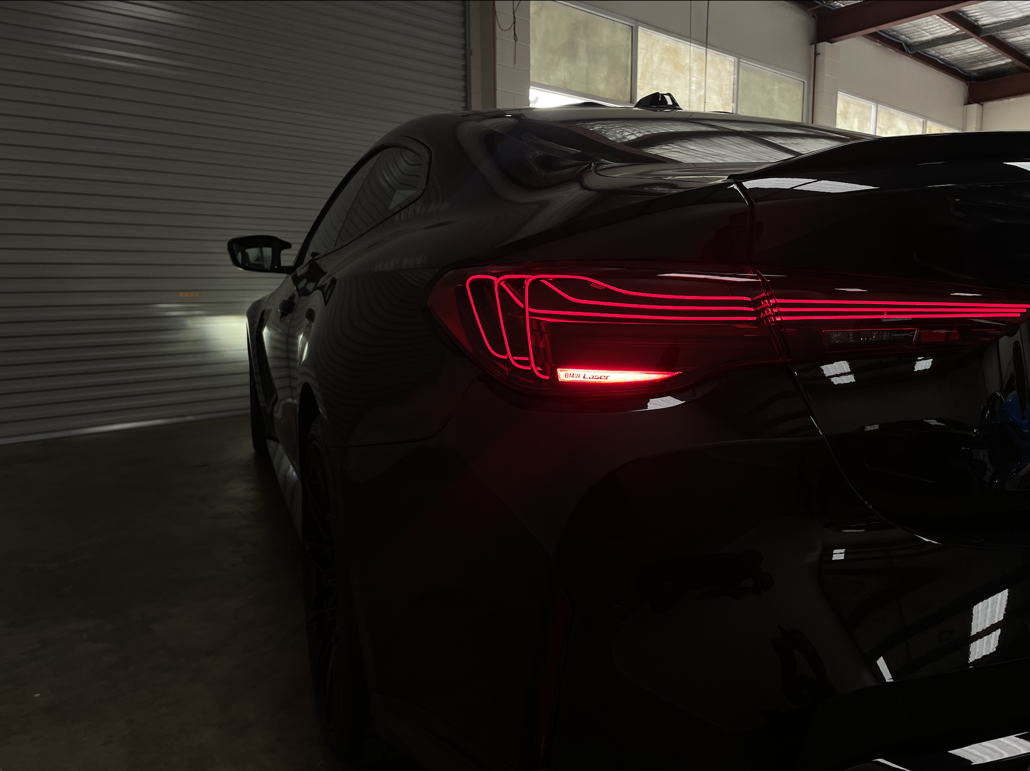 Black luxury sedan car parked inside a building with closed metal garage door, showing rear taillights and side mirror.