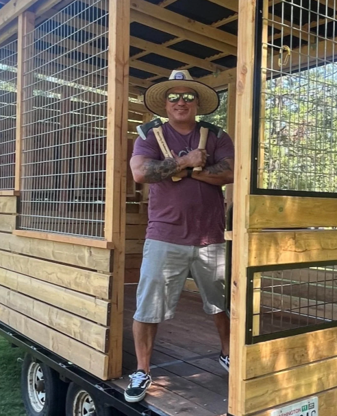Man standing inside a wooden dog kennel with wire mesh sides, wearing a straw hat, sunglasses, a purple t-shirt, gray shorts, and sneakers, smiling with arms crossed.