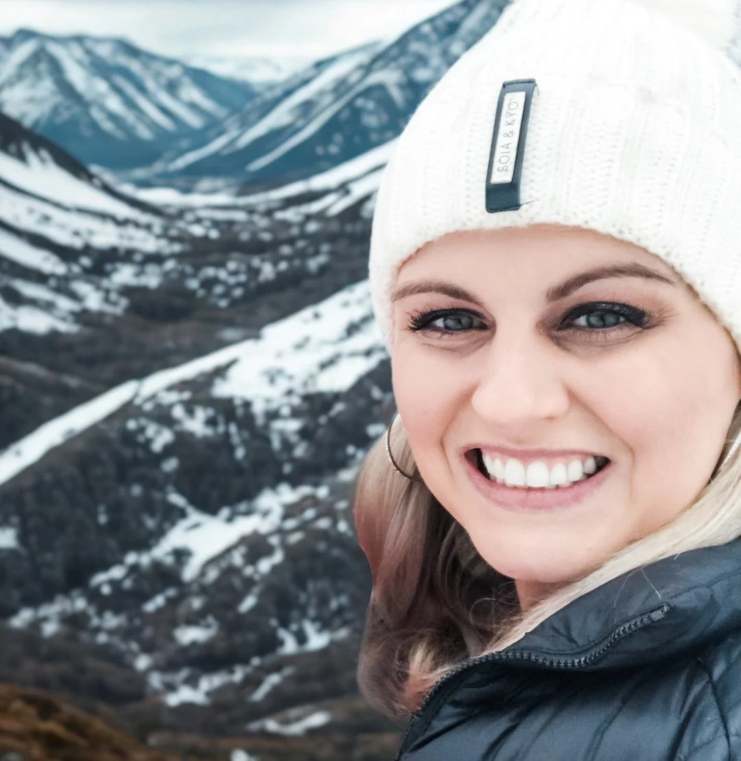 A smiling woman with blue eyes and blonde hair, wearing a white knit hat and a black jacket, stands in front of snow-covered mountains and valleys.