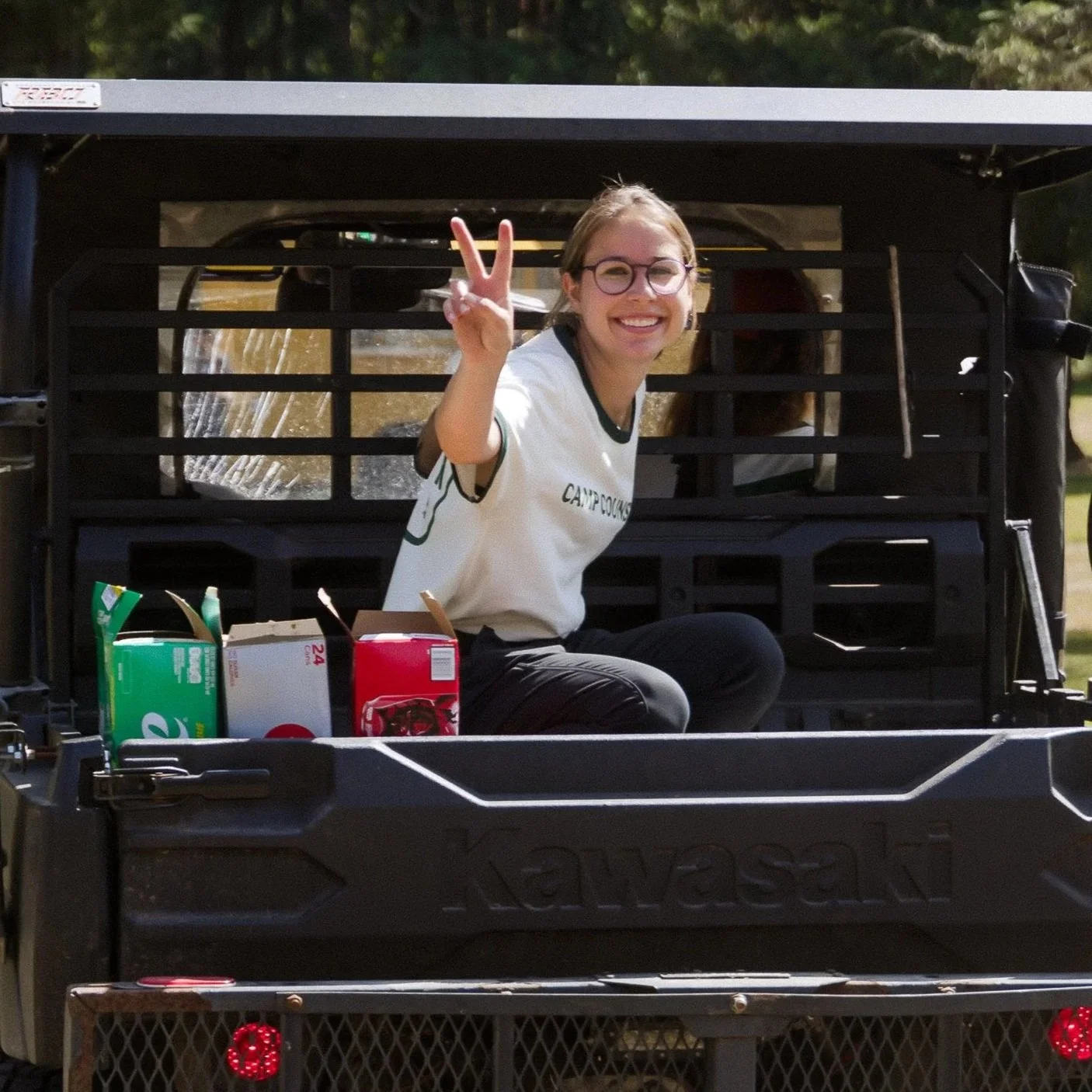 A smiling young woman with glasses making a peace sign while sitting in the back of a Kawasaki utility vehicle with boxes of tea or snacks in front of her.