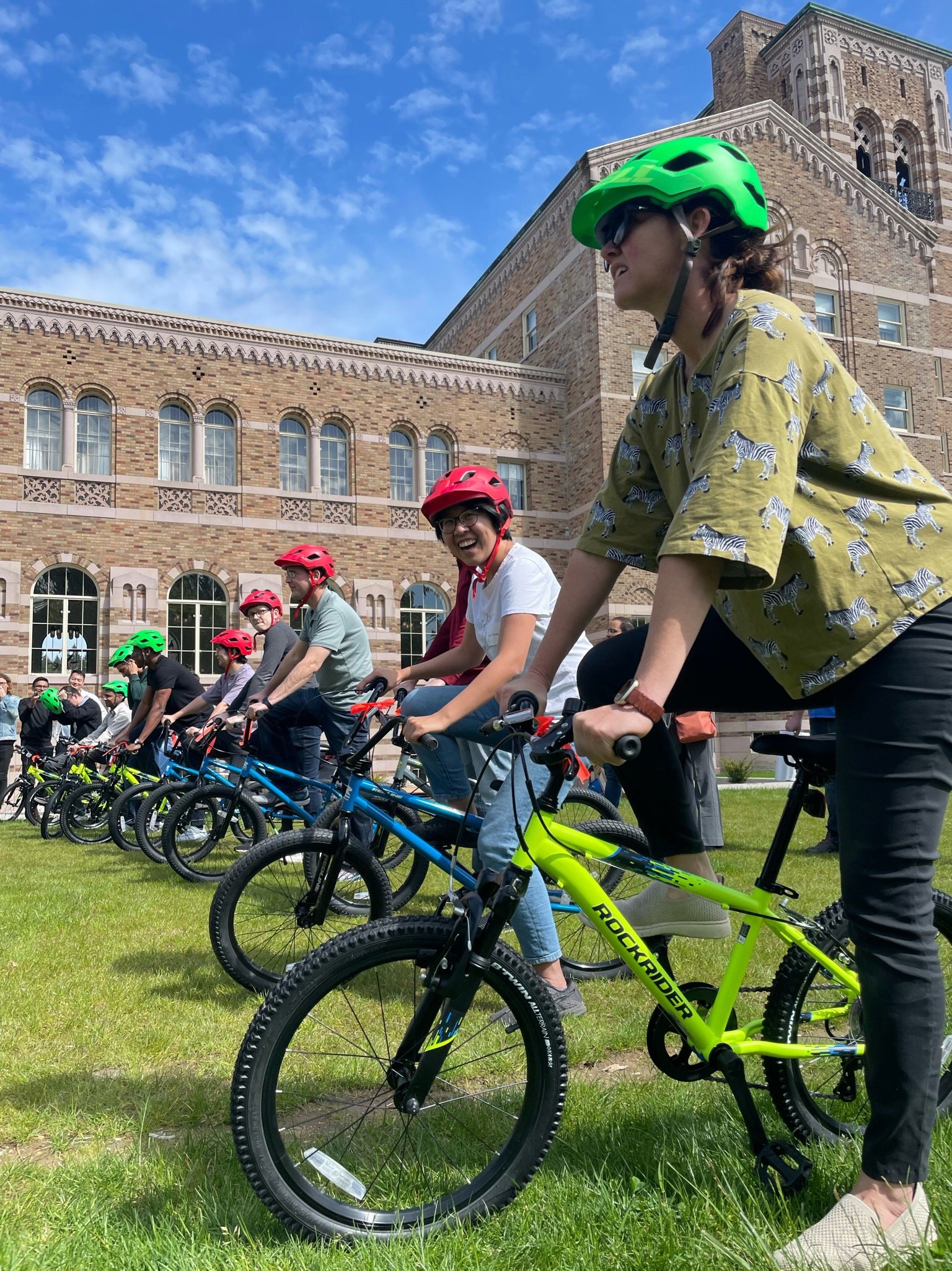 Group of people on mountain bikes, wearing helmets, gathered outdoors on grass in front of a historic brick building under a partly cloudy sky, smiling and enjoying a biking event.