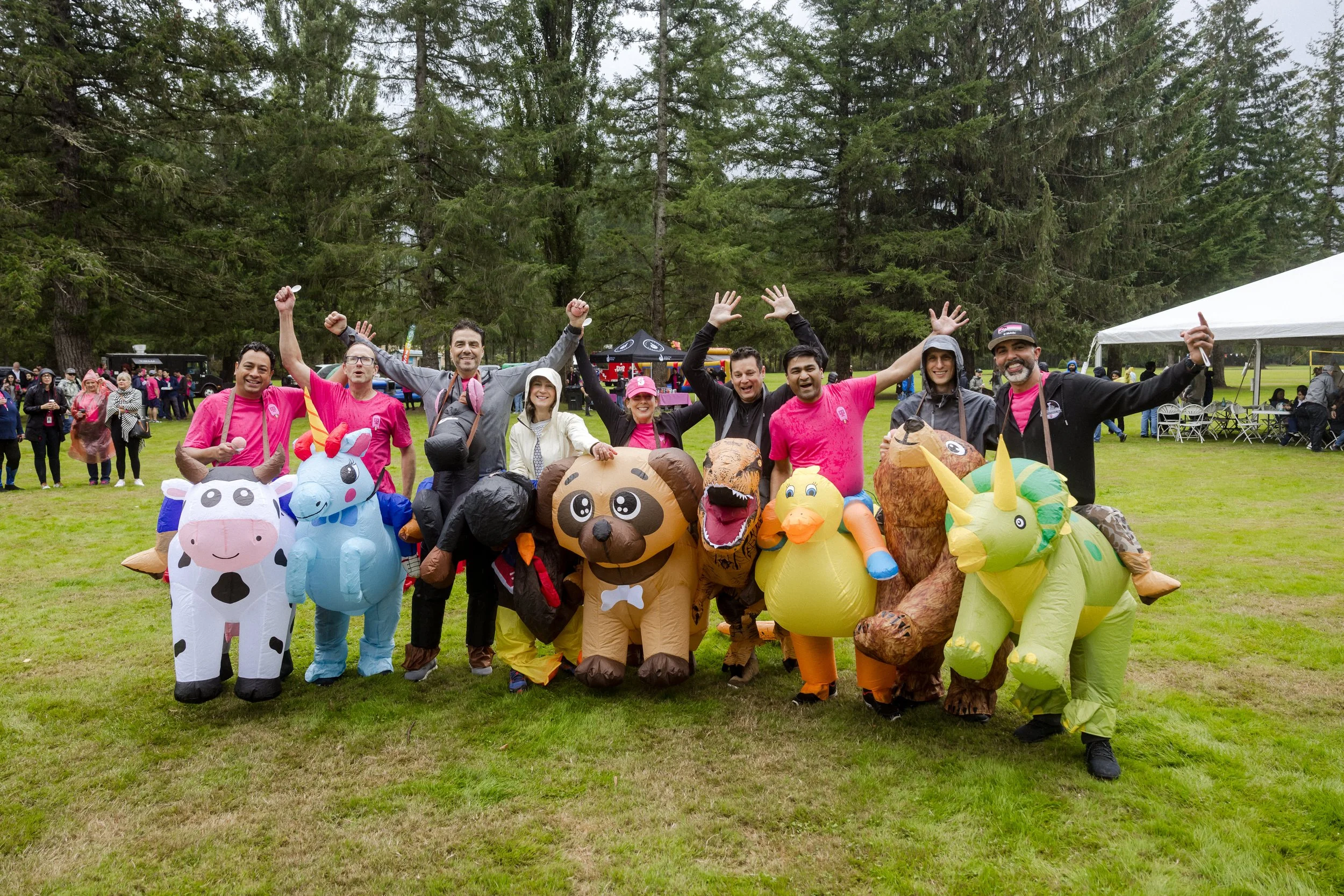 Group of people dressed in inflatable animal costumes, posing cheerfully on a grassy field with trees in the background.