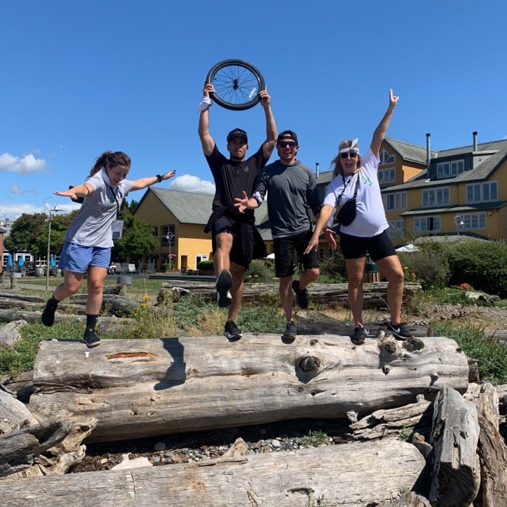 Group of four people jumping on logs outdoors on a sunny day, with buildings and a blue sky in the background.