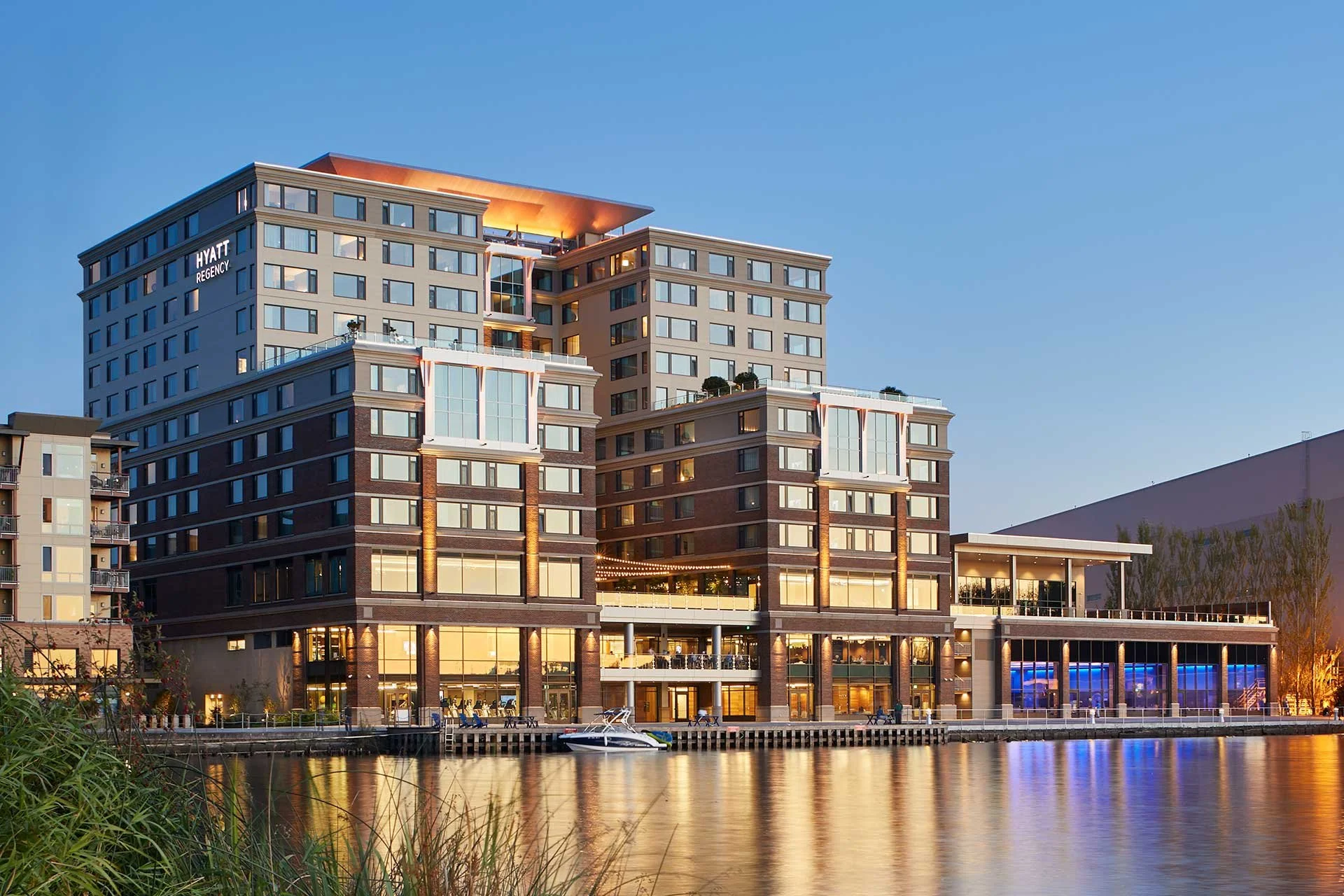 Exterior view of a modern hotel building named Hyatt Regency, situated near water, with reflections on the water surface and evening lighting.