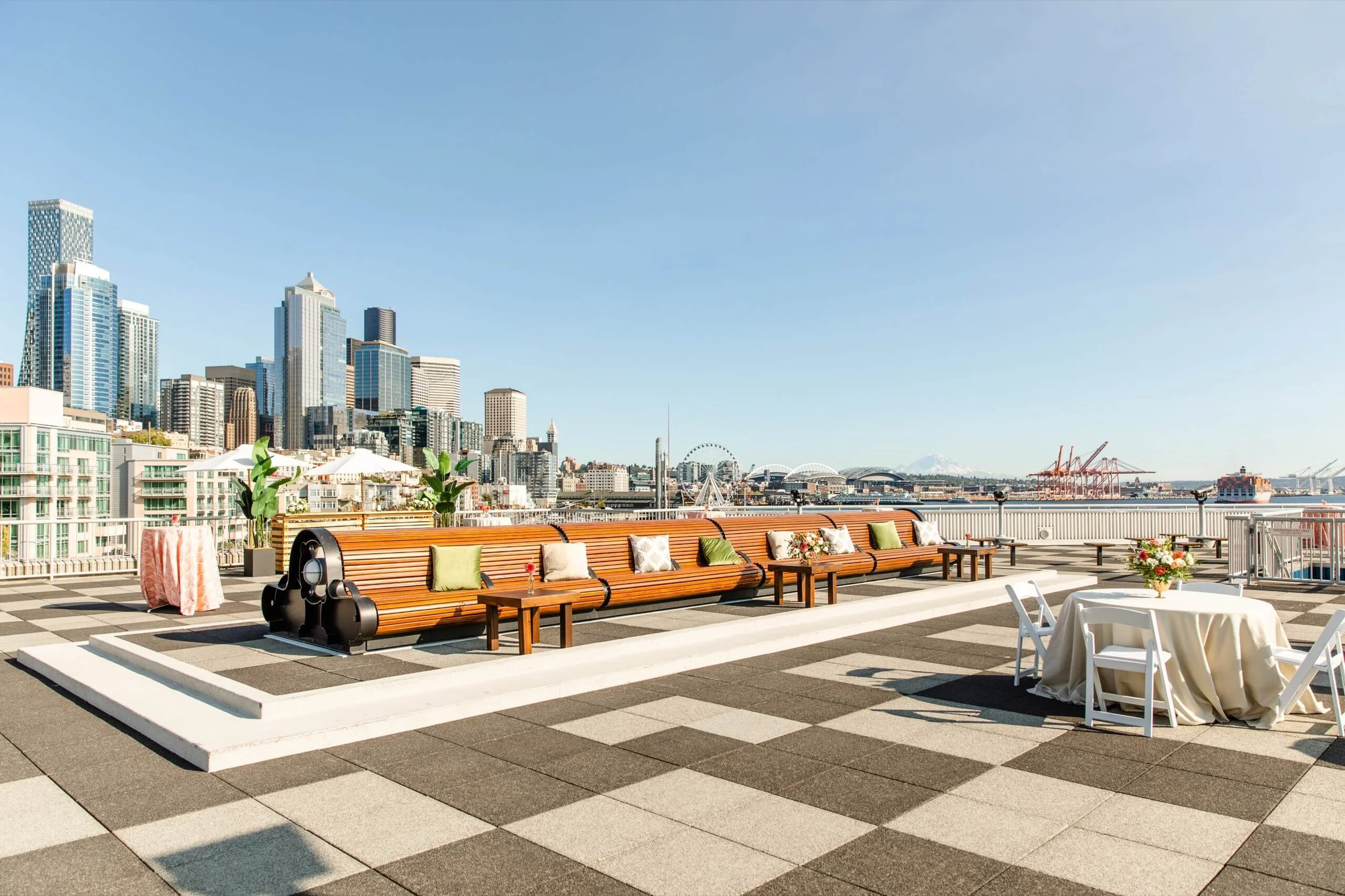 Rooftop terrace with city skyline view, featuring wooden benches with cushions, potted plants, a round table with chairs and floral centerpiece, and construction cranes and ferris wheel in the distance.